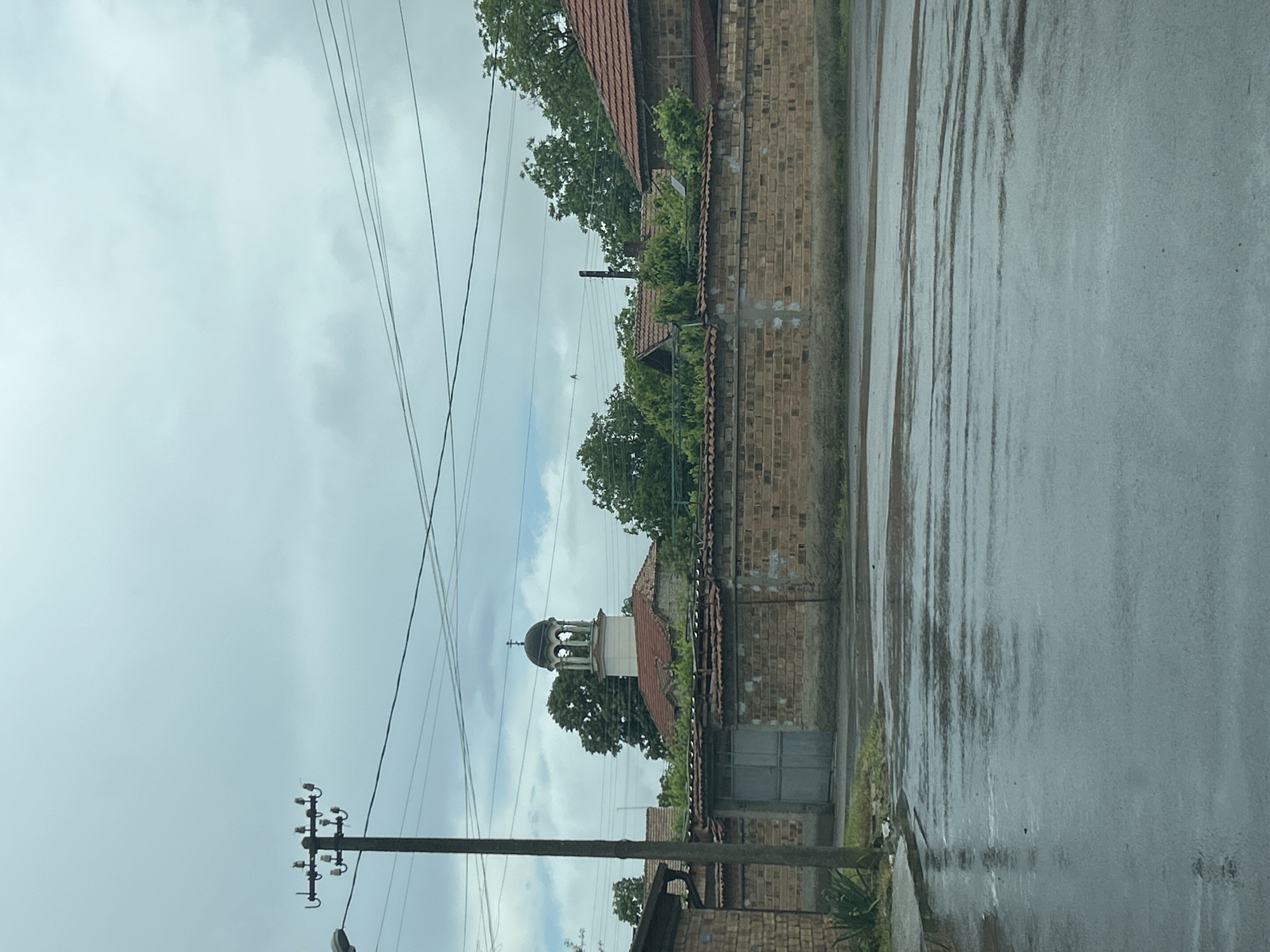 The image captures an outdoor street scene on a cloudy, overcast day, suggesting recent rain due to the visibly wet and reflective road surface.
**Subjects:** The scene features a street or road on the right, bordered by a brick wall and a narrow sidewalk on the left. Behind the wall, several buildings with red-tiled roofs are partially visible, interspersed with lush green trees. A prominent white church bell tower, topped with a dark, ornate dome and a cross, stands out against the sky, rising above the surrounding structures and foliage. In the foreground on the left, a wooden utility pole with multiple insulators supports numerous overhead power lines that stretch across the cloudy sky. A gray metal gate or door is set into the brick wall near the bottom center.
**Activity or event taking place:** No specific activity or event is captured; the image portrays a static street view. The wet ground indicates that it has recently rained.
**Notable details:** The time of day is daylight, characterized by soft, diffuse light filtering through a dense, cloudy sky. The primary weather detail is the wet condition of the street, which shows dark streaks and reflections from the water. The architectural style, including the brick wall, red-tiled roofs, and the distinctive Orthodox-style church dome and cross, provides a cultural context. The network of power lines adds an element of modern infrastructure to the otherwise traditional-looking village or town setting.
**Any visible text:** There is no visible text present in the image.