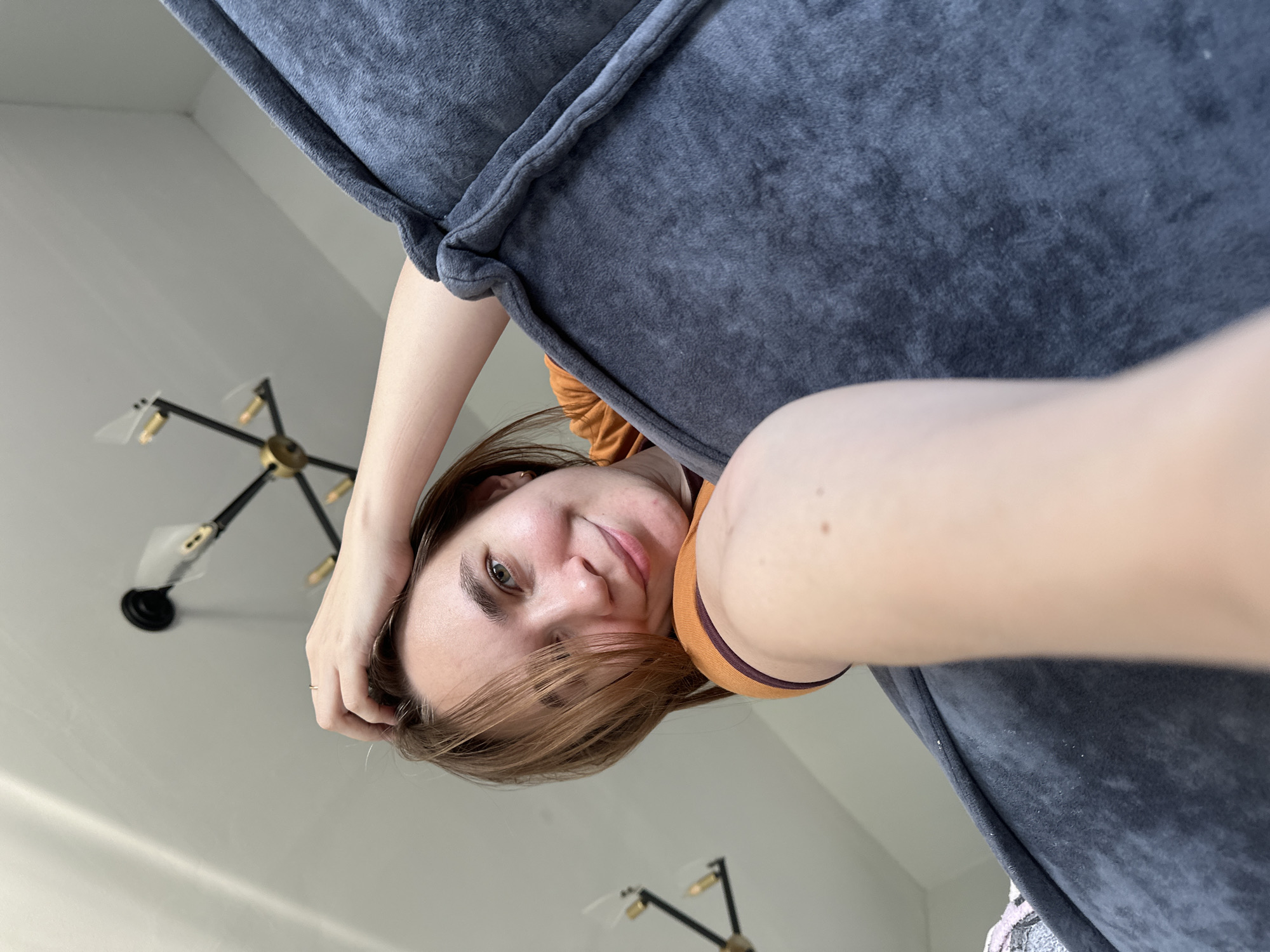 A woman with light skin and brown hair poses for a selfie. She is in an indoor environment with white walls. Part of a gray cushioned object, possibly a sofa, is visible. A modern chandelier with black and gold fixtures is attached to the ceiling. There is no verifiable evidence to identify this location as Kharnavy, Russia.