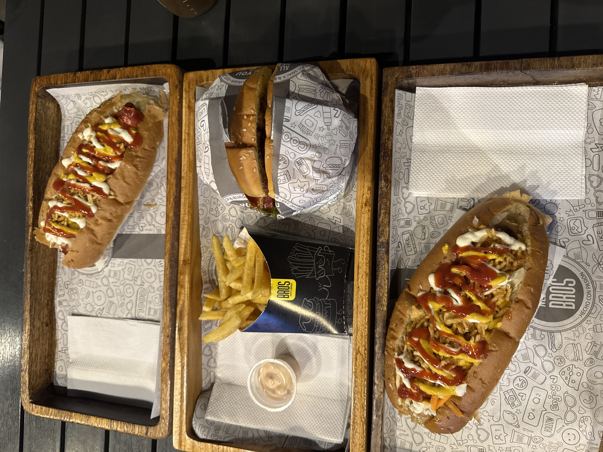 The image shows three wooden trays of food on a dark, slatted table. The trays each contain branded paper liners with cartoon drawings and the word "BROS" repeated. The tray on the left contains a long sandwich topped with a yellow sauce, a red sauce, and small fried potatoes. The middle tray has a hamburger wrapped in similar branded paper, along with a container of french fries in a black "BROS" branded box, and a small container of sauce. The tray on the right has a paper napkin and a similar sandwich to the one on the left, also topped with sauces and fried potatoes. Based on the branding "BROS" on the food containers, and the reference to "HECHO CON PASION," the food is likely from a "BROS" location. The trays and branding suggest a fast-casual dining environment. The presence of Spanish text suggests this is in a Spanish-speaking location, potentially Venezuela.