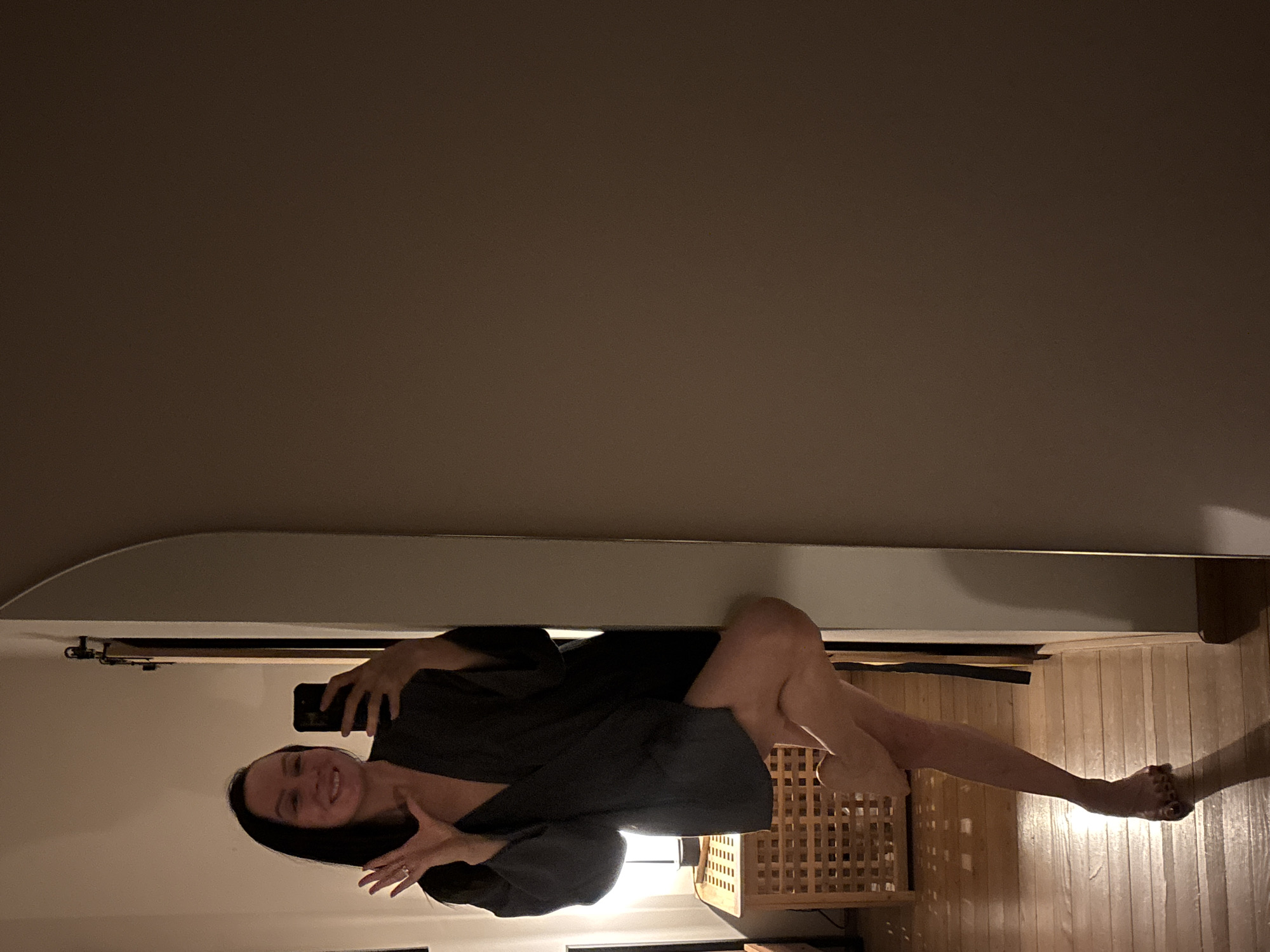 The image shows a woman lying on her side in what appears to be an indoor setting. She is holding a smartphone and facing the camera. The background includes a bedframe, a wooden lattice-like object, and a wooden floor. The location is not discernible from the image.
