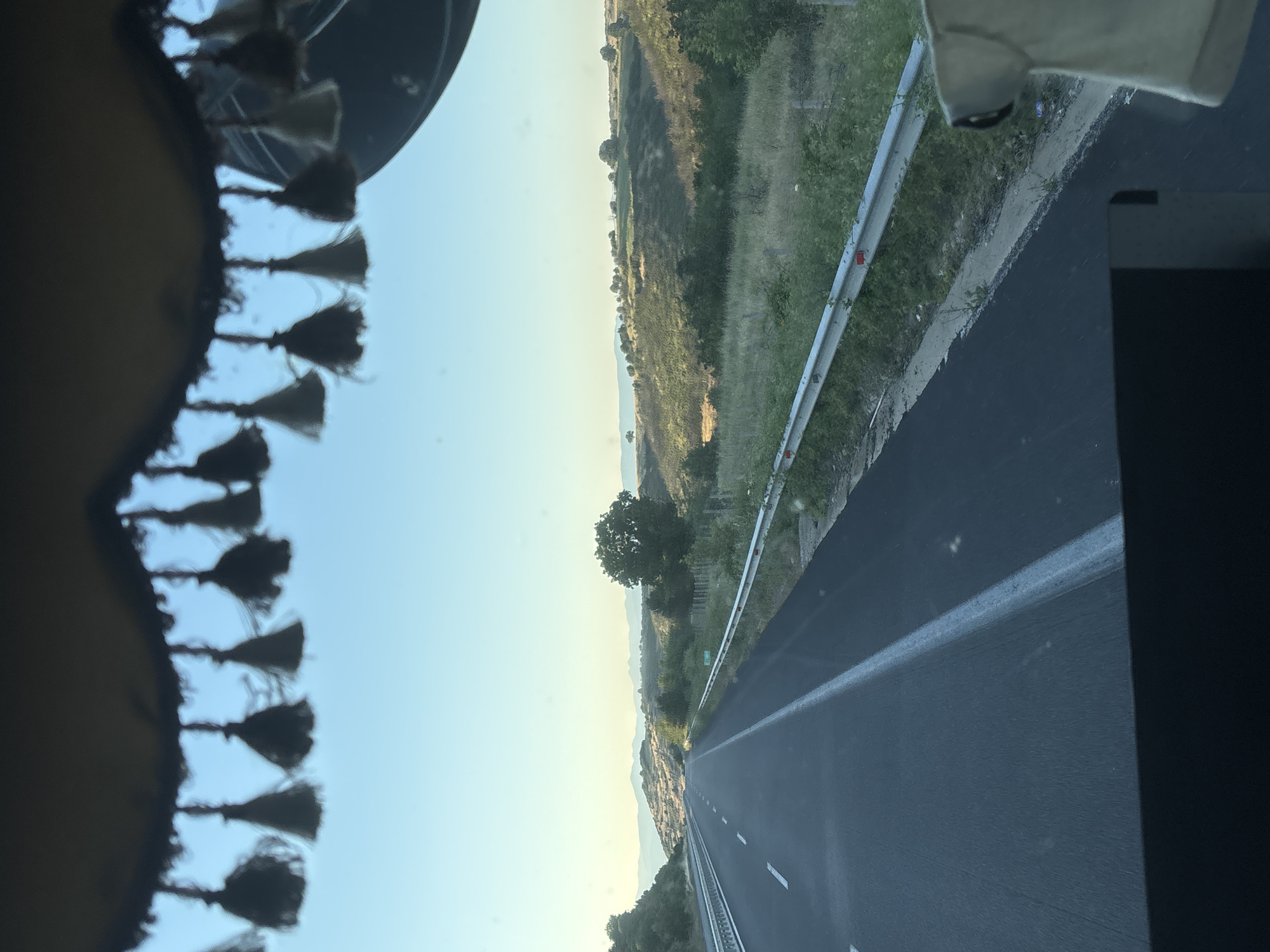 The image shows a view from inside a vehicle looking out onto a highway in Avra, Greece. The highway stretches into the distance, with rolling hills and sparse vegetation on either side. The sky is clear and blue. A metal guardrail runs along the right side of the highway. The top of the image shows a fringe of decorative tassels hanging from the vehicle's interior. There are no people visible in the image.