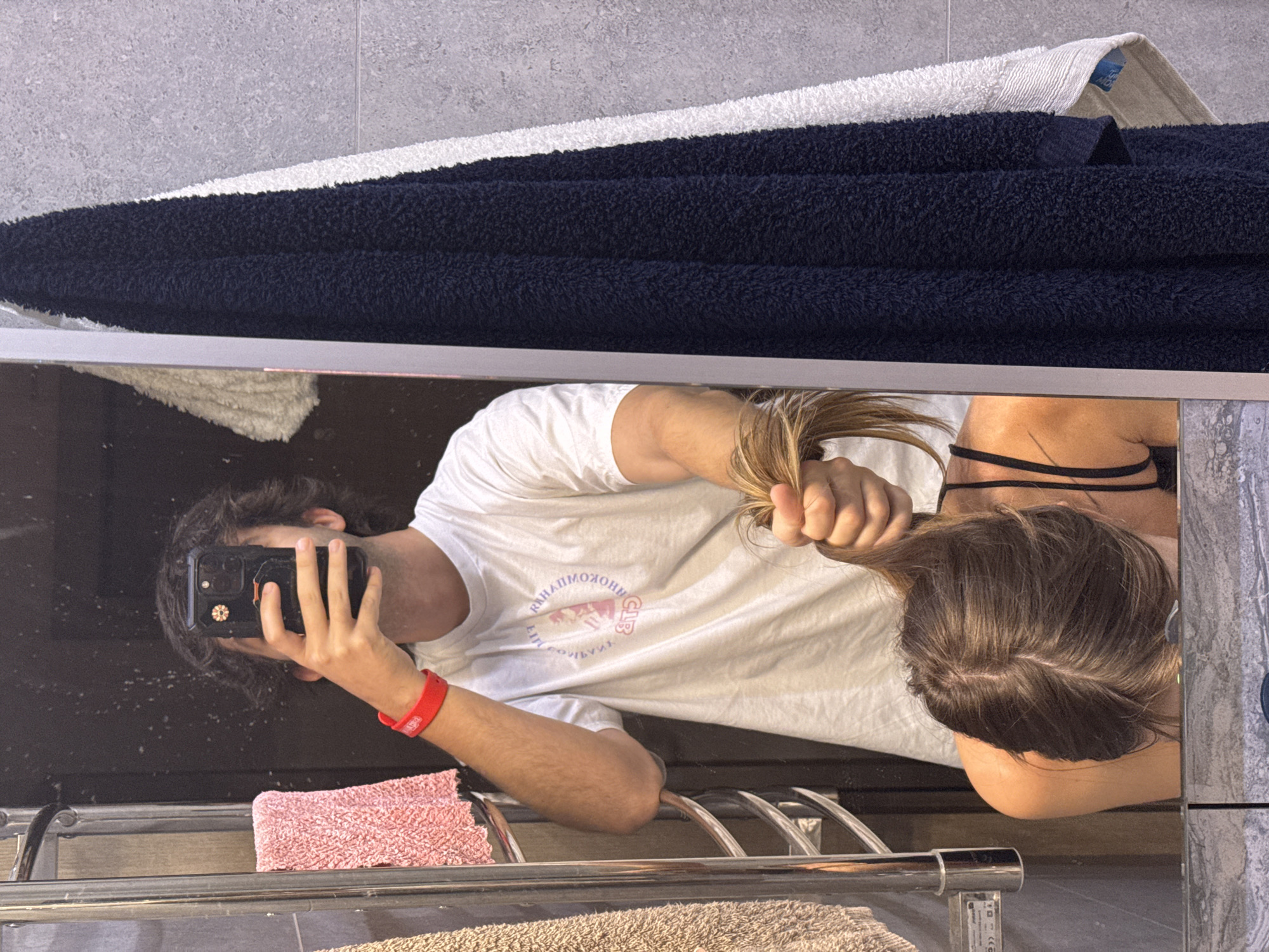 In a bathroom, a young person with dark, messy hair and a red wristband is taking a mirror selfie with their phone. They are wearing a white t-shirt with a circular graphic. Their left hand is holding the phone, and their right hand is pulling a clump of blonde hair. In the mirror's reflection, another person's head is visible, bent forward, with their hair being pulled. The bathroom features a silver heated towel rack with a pink towel and a beige towel. Dark navy and white towels hang on the tiled wall. The scene is well-lit, and the camera appears to be at eye level. The location is not specified.
