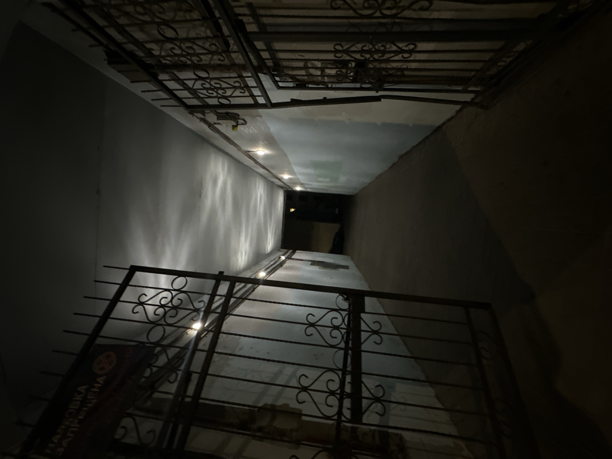 This image depicts a long, narrow, dimly lit covered passageway or corridor in the city of Unknown, Unknown. The perspective is directed down the length of the passage. Two dark metal gates, each featuring ornate scrollwork patterns, stand open on either side of the foreground, leading into the corridor. A rectangular sign, dark blue with white Cyrillic text reading "ПАРКОВКА ЗАПРЕЩЕНА" (Parking Forbidden) and a red 'no parking' symbol, is affixed to the left gate.
The walls of the corridor are painted, appearing light blue or off-white on the upper section and a darker shade on the lower half, exhibiting some wear and discoloration. Exposed electrical conduits or wiring are visible along the upper right wall. Multiple bare light bulbs are mounted along the ceiling on both sides of the corridor, casting reflections on the off-white, seemingly textured ceiling and providing the primary source of illumination. The overall lighting is dim, creating deep shadows. The floor is a dark, uniform surface, likely concrete or asphalt. A faint light source is discernible at the far end of the corridor, indicating a distant exit or another illuminated area. No individuals or active subjects are present within the scene.