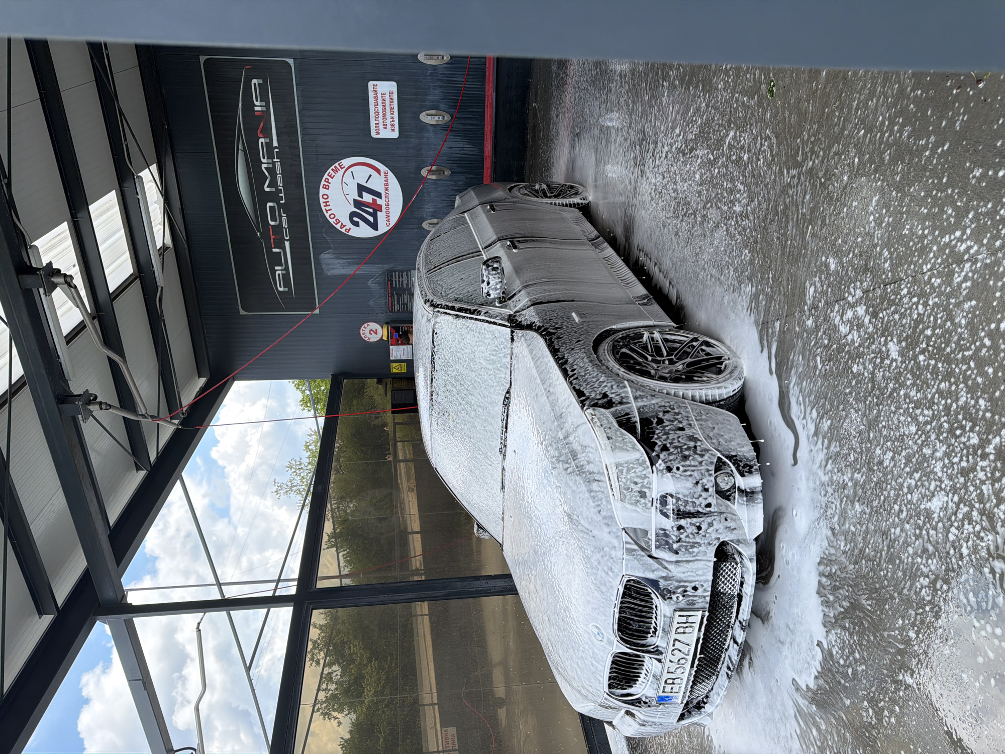 The image depicts a silver BMW car undergoing a car wash at "Auto Mania Car Wash," which offers 24/7 self-service. The license plate of the car reads "EB5627BH," presumably issued in Bulgaria. The car is covered in soap foam, indicating it's in the washing process. The car wash bay has a metal structure with a partial glass enclosure. Visible signage indicates "РАБОТНО ВРЕМЕ 24/7" (Working Hours 24/7) and instructions to dry the vehicles outside the bays. The location is inferred to be Gabrovo, Bulgaria, based on the license plate and the presence of Bulgarian text.