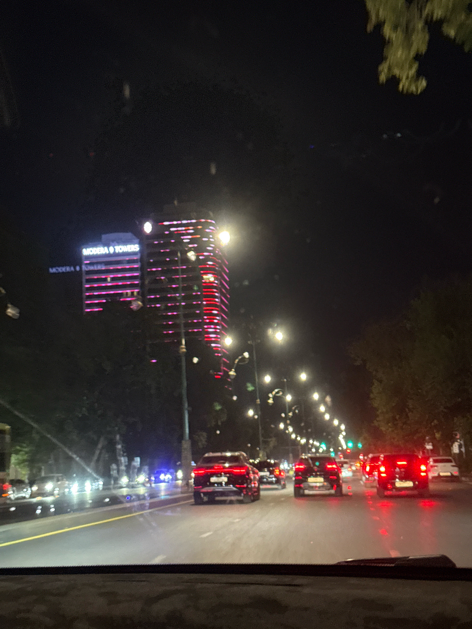 The image depicts a night-time street scene in Tashkent, Uzbekistan, on the avenue near the "Modena Towers". Several cars are visible on a multi-lane road, with their headlights and taillights illuminated. In the background, the "Modena Towers" building complex is prominently lit with pink and red lights, indicating it is a commercial or residential area. Streetlights line the road, casting a glow on the wet asphalt, suggesting recent rain or that the road is being watered. Trees are visible on the sides of the road, with some branches extending over the street. The perspective is from inside a vehicle, looking forward through the windshield, which has a dashboard or wiper visible in the foreground. The overall atmosphere is that of a busy urban environment at night, with the architectural lighting of the towers being a significant focal point.