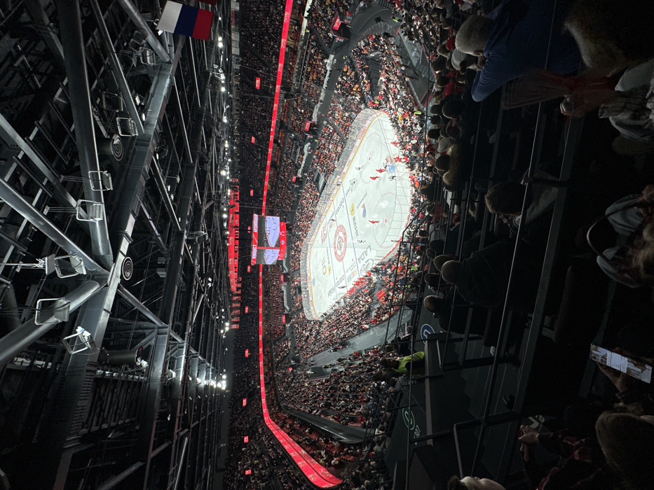 The image displays an indoor ice hockey game in progress at the Uralets Arena in Yekaterinburg, Russia. The perspective is an elevated view from the upper seating bowl, looking down onto the ice rink. Thousands of spectators occupy the dark-colored stadium seats, predominantly clad in dark clothing, with pockets of red visible in areas.
The ice rink, centrally located and brightly illuminated, features hockey players in red and white uniforms actively engaged in the game. Prominent sponsor logos such as "LADA" and a circular team emblem are visible on the ice surface. A large central scoreboard above the rink displays live game action, "ВИКИНГ," and the team name "АВТОМОБИЛИСТ" (Avtomobilist). Surrounding the seating tiers are illuminated red digital advertising boards and static red banners.
The arena's ceiling consists of a complex network of dark grey structural beams, ventilation ducts, and various lighting fixtures. A Russian flag, with its white, blue, and red horizontal stripes, hangs from the ceiling structure in the upper right portion of the frame. In the immediate foreground, several spectators are partially visible; one individual on the far left holds a smartphone, its screen displaying images. The overall environment is a large, enclosed sports venue characterized by high-contrast lighting: brilliant white light on the ice, darker ambient light in the stands, and striking red accent lighting throughout the arena.