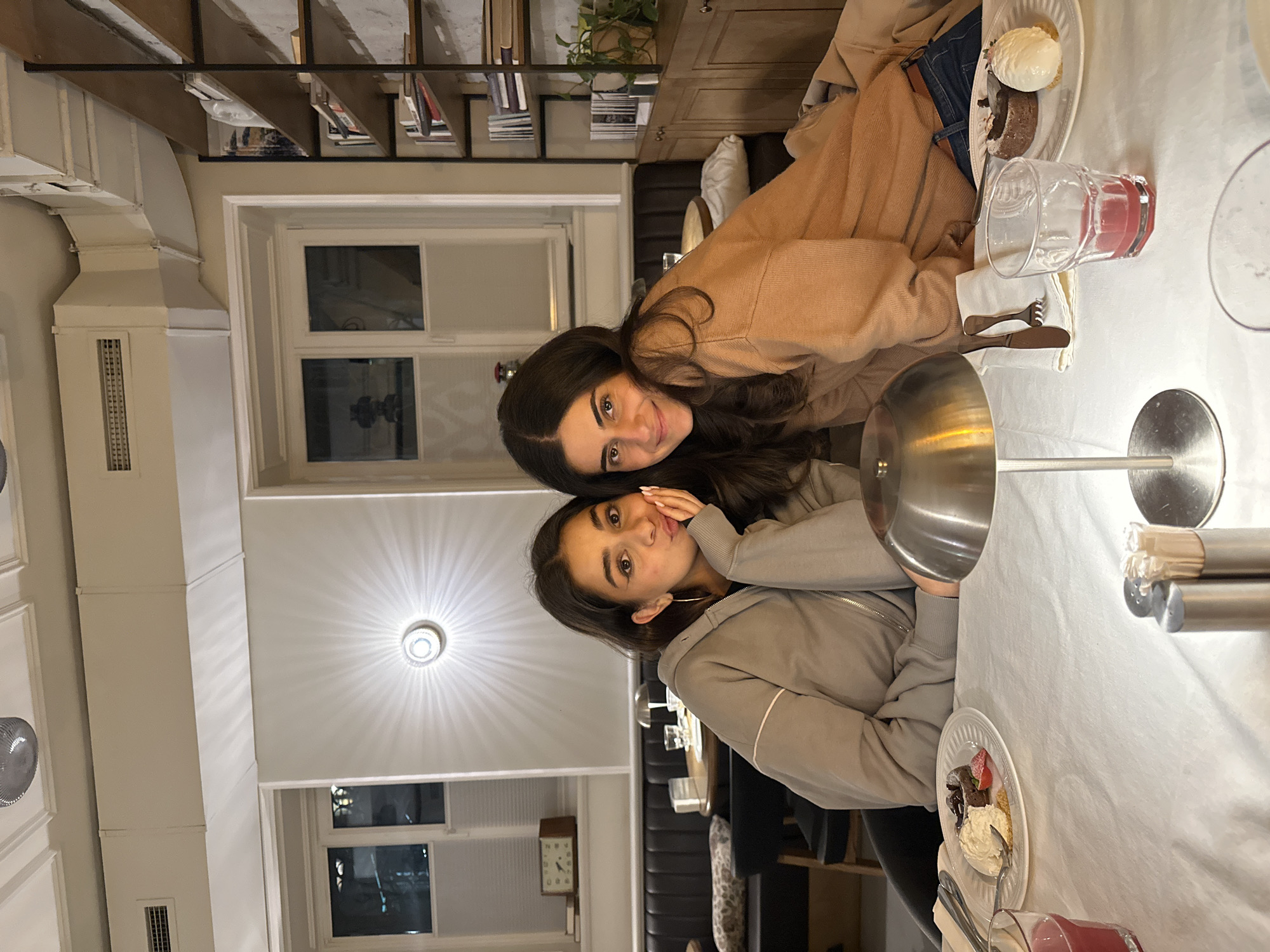 Two women pose for a photograph while sitting at a table in what appears to be an indoor setting. Both are smiling and looking at the camera. The woman closer to the camera is wearing a gray zip-up hoodie. The other woman is wearing a tan sweater.
The table is covered with a white tablecloth and has plates with desserts and drinks. One plate has what looks like a scoop of ice cream and a piece of cake. There are two glasses filled with red liquids on the table. Silver table lamps are also visible.
The background includes a bookcase filled with books, windows, a decorative light fixture on the ceiling, and architectural details, including ductwork and paneling.
Without additional location cues it is impossible to verify if this is Yerevan. The details provided are limited to the visual components of the image.