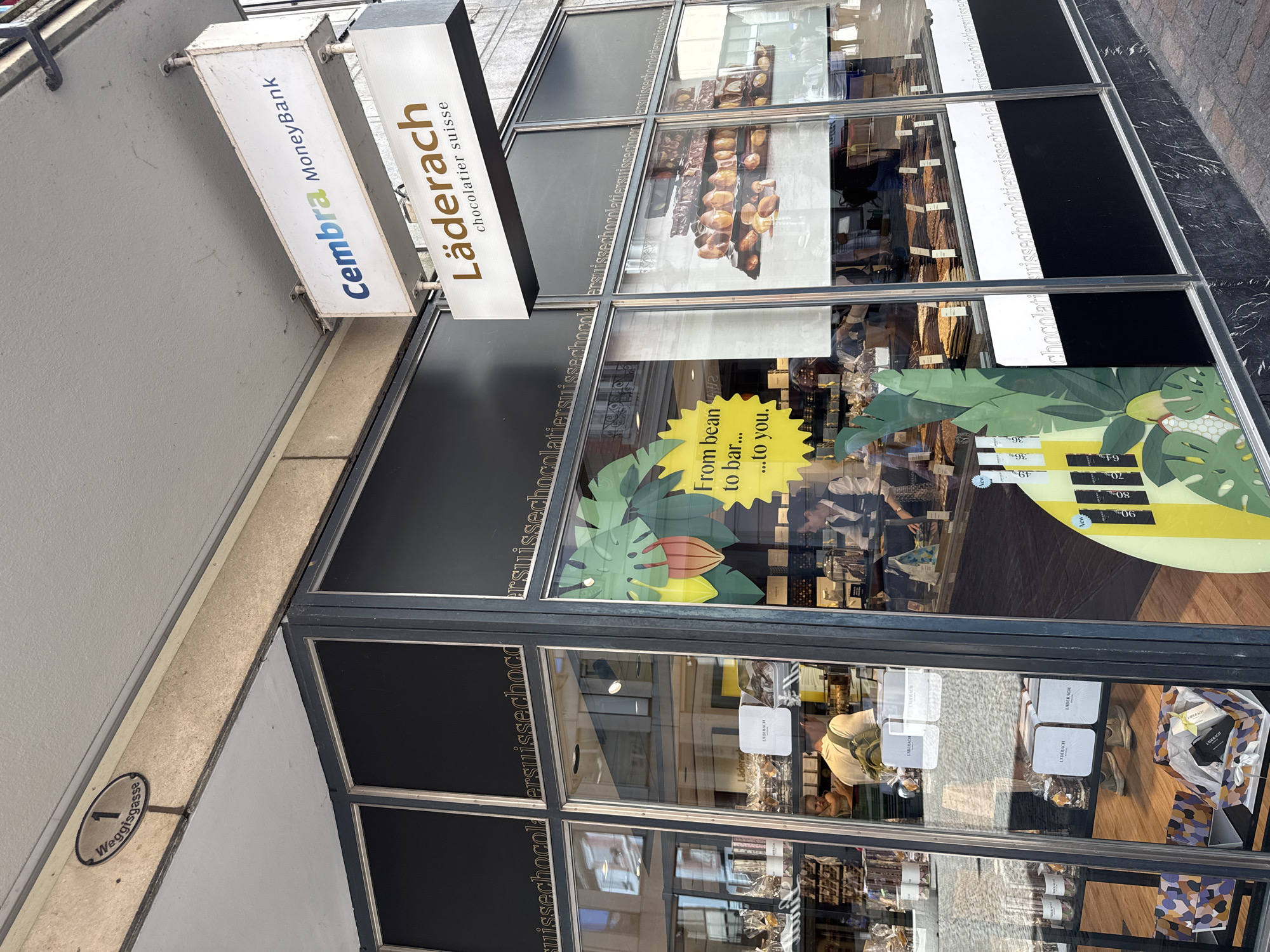 This is a vertical, eye-level, outdoor photograph of the facade of a Chocolatier Suisse store in Lucerne, Switzerland. The store features large display windows showcasing its products and interior. A prominent sign above the entrance reads "Läderach chocolatier suisse" in gold lettering on a dark background, with another smaller sign for "Cembra MoneyBank" to the left.
The display windows are filled with various chocolate bars, boxes, and displays of cocoa beans, suggesting an active retail environment. One window has a large graphic featuring tropical leaves, a cocoa pod, and a yellow banner with text including "From bean to bar... to you." Prices for chocolate bars are displayed on a yellow background with labels like "36%", "49", "64", "70", and "90".
Reflections in the glass reveal the street scene outside, including buildings and what appears to be a person walking. Inside the store, a person is visible in the background, appearing to be a staff member or customer. The overall impression is of a high-end chocolate shop in a commercial area, likely during daylight hours. Street signage for "Weggisgasse" is visible on the lower left of the image.