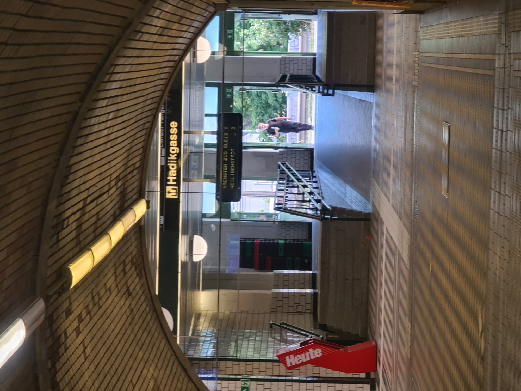 The image shows an interior view of a Vienna U-Bahn station. A sign above an archway reads "Hadikgasse." In the background, a man walks towards the camera holding a bottle. The station features tiled walls, a wooden arched ceiling, and illuminated signs. A red newspaper stand is visible on the left side of the image. The floor is tiled with a patterned surface, indicating a public transit space.