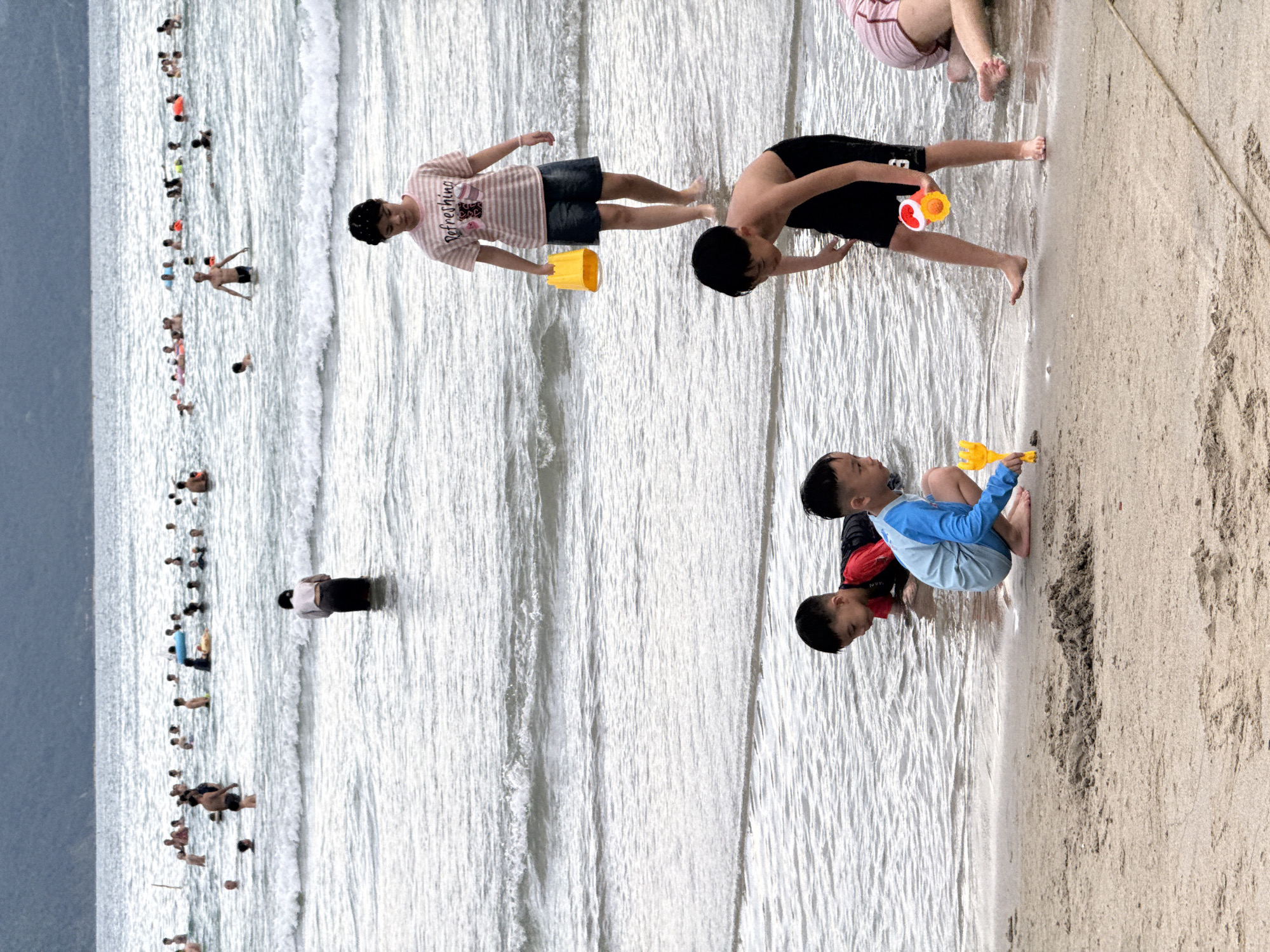 This media file captures a beach scene in Đà Nẵng, Vietnam, likely during daylight hours given the bright, reflective water and sandy beach. The water is moderately wavy, suggesting a pleasant day for beach activities.
The primary subjects are numerous people enjoying the ocean and the shoreline. In the foreground, two young boys are visible. One is wearing a blue and light blue rash guard and appears to be playing with a yellow toy. The other boy, partially obscured, is wearing a red rash guard. Further back, a person in a striped t-shirt and denim shorts stands in the water holding a yellow bucket. Another person is seen in dark shorts and a dark top, also in the water. In the distance, a long line of people are wading or swimming in the ocean, indicating a popular beach day. A single person stands further out in the water, dressed in dark shorts and a light-colored top.
The setting is clearly a beach with sand and ocean. The water is a light blue-grey, reflecting the sky, and the sand is a light tan. The overall mood appears to be one of leisure and enjoyment, with people engaging in typical beach activities like swimming and playing. There are no visible textual elements in the image.