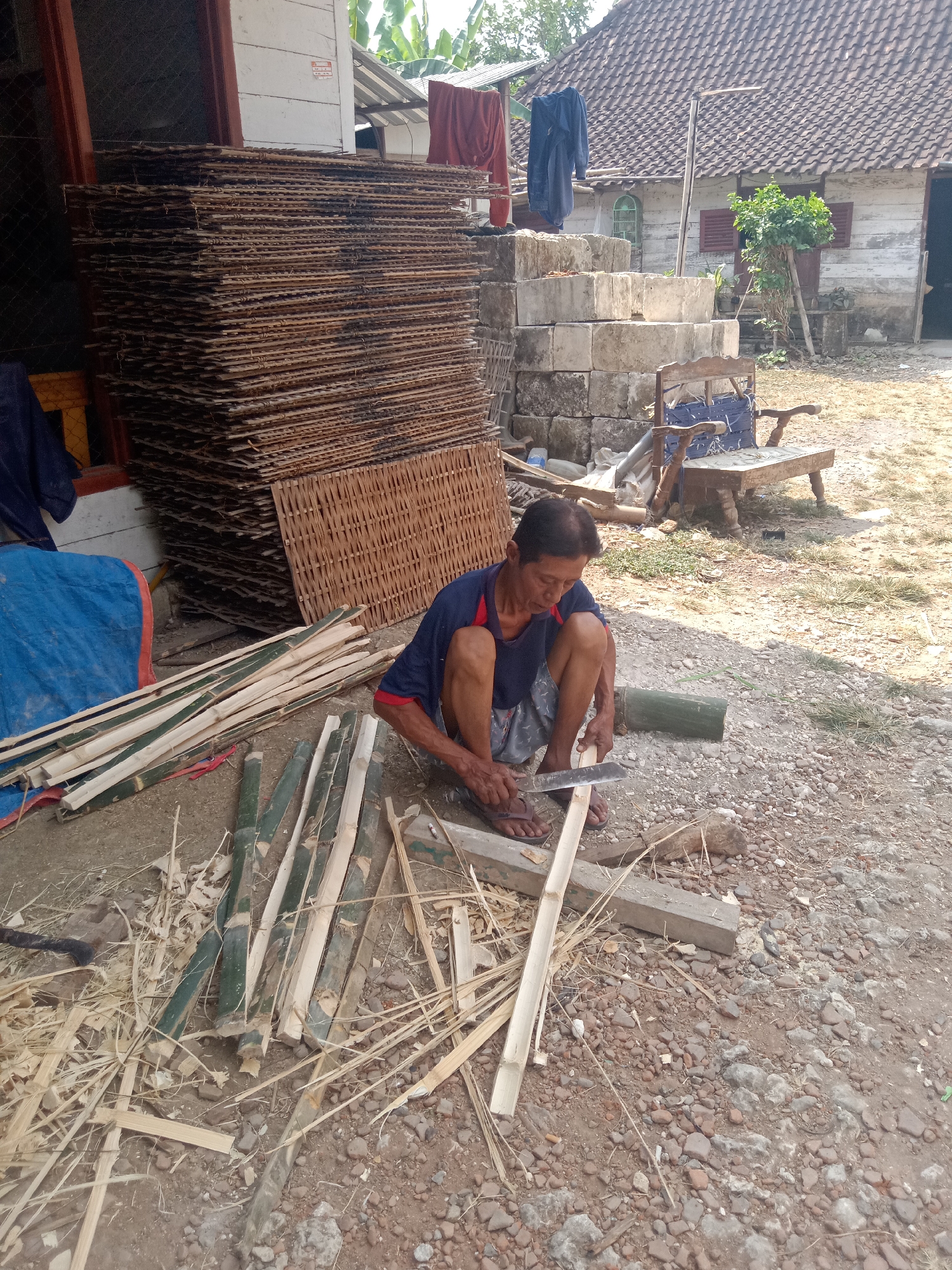 In Jumput, Indonesia, an outdoor scene depicts a man of Asian descent crouched on the ground, meticulously splitting bamboo poles with a large knife. He is dressed in a blue shirt with red trim and patterned shorts. The ground is covered in dirt, gravel, and scattered bamboo debris. In the background, a stack of woven mats leans against a building with a white facade and a tiled roof. Adjacent to the mats are several large concrete blocks, and further back, a rustic wooden bench sits under the shade of a tree. The overall environment suggests a rural or workshop setting.