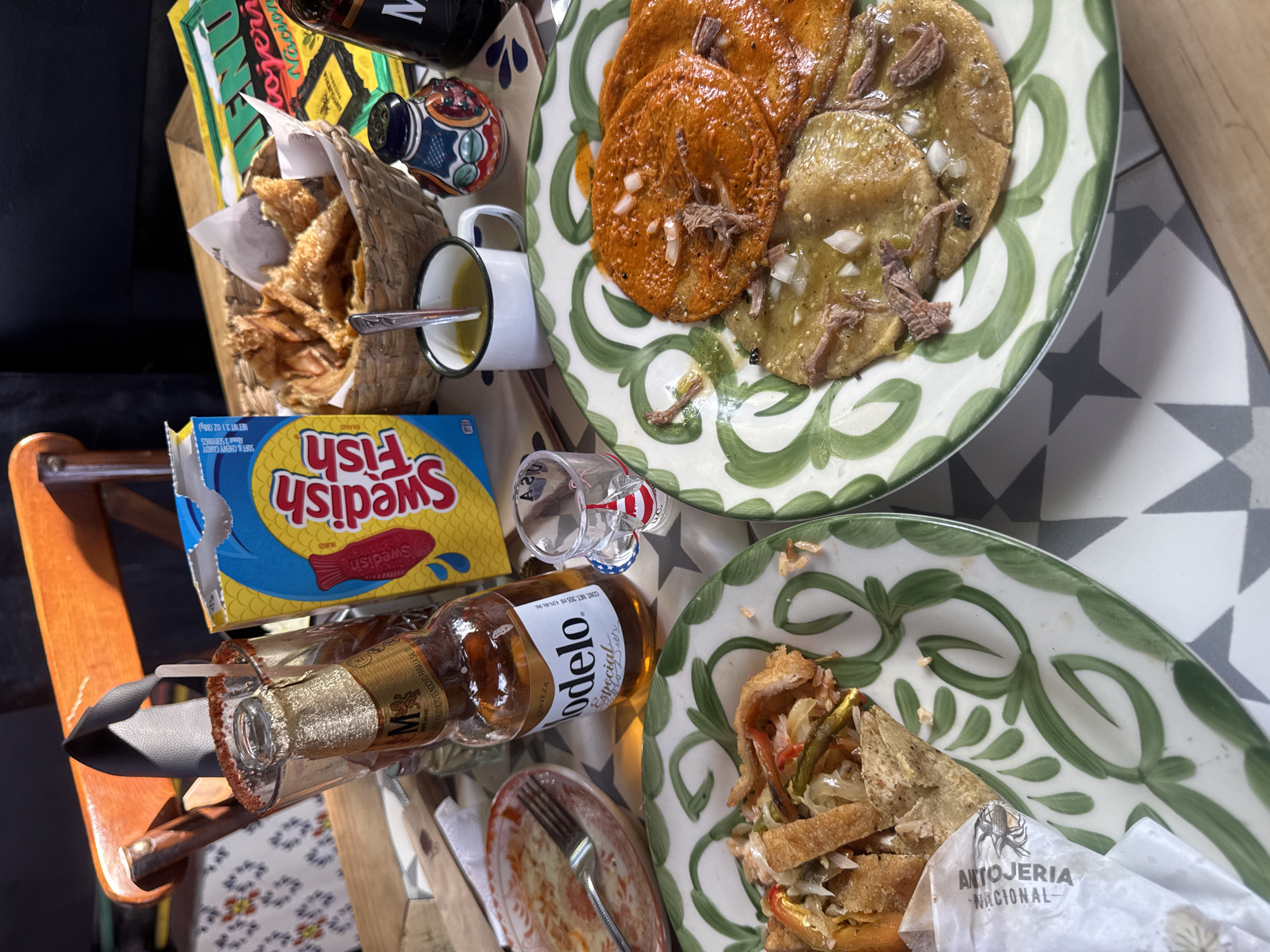 This media file captures an overhead view of a casual meal spread on a table, likely at an antojeria in Puebla, Mexico, given the local context and dishes.
The main subjects are various food items and beverages arranged on a patterned tabletop. In the upper right, a plate features two types of antojitos: several orange/red, round, flat masa preparations (possibly sopes or enchiladas) and two green ones, all topped with shredded meat and diced white onion. Below this, another plate holds what appears to be a fried torta or similar sandwich-like dish, filled with shredded meat, vegetables, and peppers, wrapped partially in paper. A woven basket contains golden-brown, crispy fried snacks, possibly chicharrones or totopos.
Beverages include a bottle of Modelo Especial beer lying horizontally, with its rim coated in chili-lime seasoning, and a clear shot glass also with a similar rim. A white enamel mug with a spoon holds a light-colored liquid. A blue box of "Swedish Fish" brand soft & chewy candy is also visible. Other objects include a colorful ceramic condiment shaker and a partially visible menu or magazine.
The scene depicts a vibrant and hearty meal. The bright lighting suggests it is daytime. The patterned floor tiles and the style of the dishes contribute to a Mexican culinary atmosphere.
Visible text includes:
- On the paper wrapping/napkin: "ANTOJERIA NACIONAL-"
- On the beer bottle: "Modelo Especial Cerveza," "CONT. NET. 355 ml 4.5% Alc Vol.," and "ESTE PRODUCTO ES NOCIVO PARA LA SALUD." The letter "M" is also visible.
- On the candy box: "Swedish Fish BRAND," "SOFT & CHEWY CANDY," "About 3 SERVINGS," and "NET WT 3.1 OZ (88g)."
- On the menu/magazine: Partially visible words like "Nacional," "cojeri," "ENT," and "D.C."