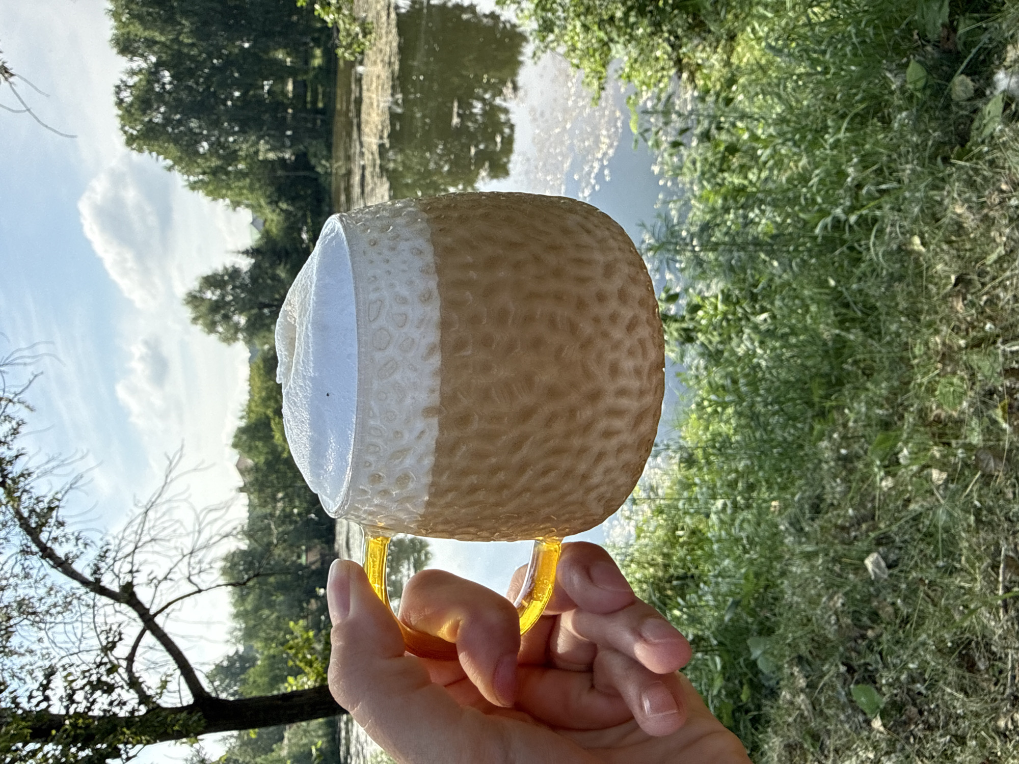 A person's hand, with visible fingernails, holds a textured, glass mug filled with a light brown beverage topped with white foam. The mug has a bright yellow handle. The background features a calm body of water, likely a river or lake, reflecting the sky and trees. Lush green foliage and trees are present on the banks of the water. The sky is blue with scattered white clouds. The scene appears to be an outdoor setting, possibly a park or natural area, in or near the city of Chaikovsky, Russia.