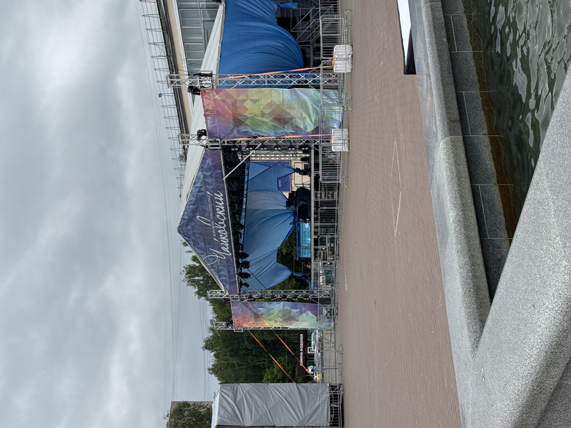 The image shows an outdoor stage setup with scaffolding and large banners. The stage is covered by a blue tent-like structure, and a large, colorful banner with a geometric pattern is suspended in front of it. Another banner with similar geometric patterns and text is visible at the bottom of the stage. A partial view of a building or structure is present in the background, and trees are visible behind the stage. A stone-tiled water feature or fountain is to the right of the frame. The weather appears overcast, suggesting a cloudy day. The overall scene depicts preparations for an outdoor event, possibly a festival or concert, in a public space.
Visible text includes "Чайковский" (Chaikovsky) and "ГОРОД" (City) on one of the banners, indicating the location. Another banner at the bottom has the text "СЕМЕНА-ВОДОЛЕЙ" (Semen-Vodoley). A small portion of text that reads "ЕРЖНСКОГО КРАЯ" (Pershinskogo Kraya) is also visible.