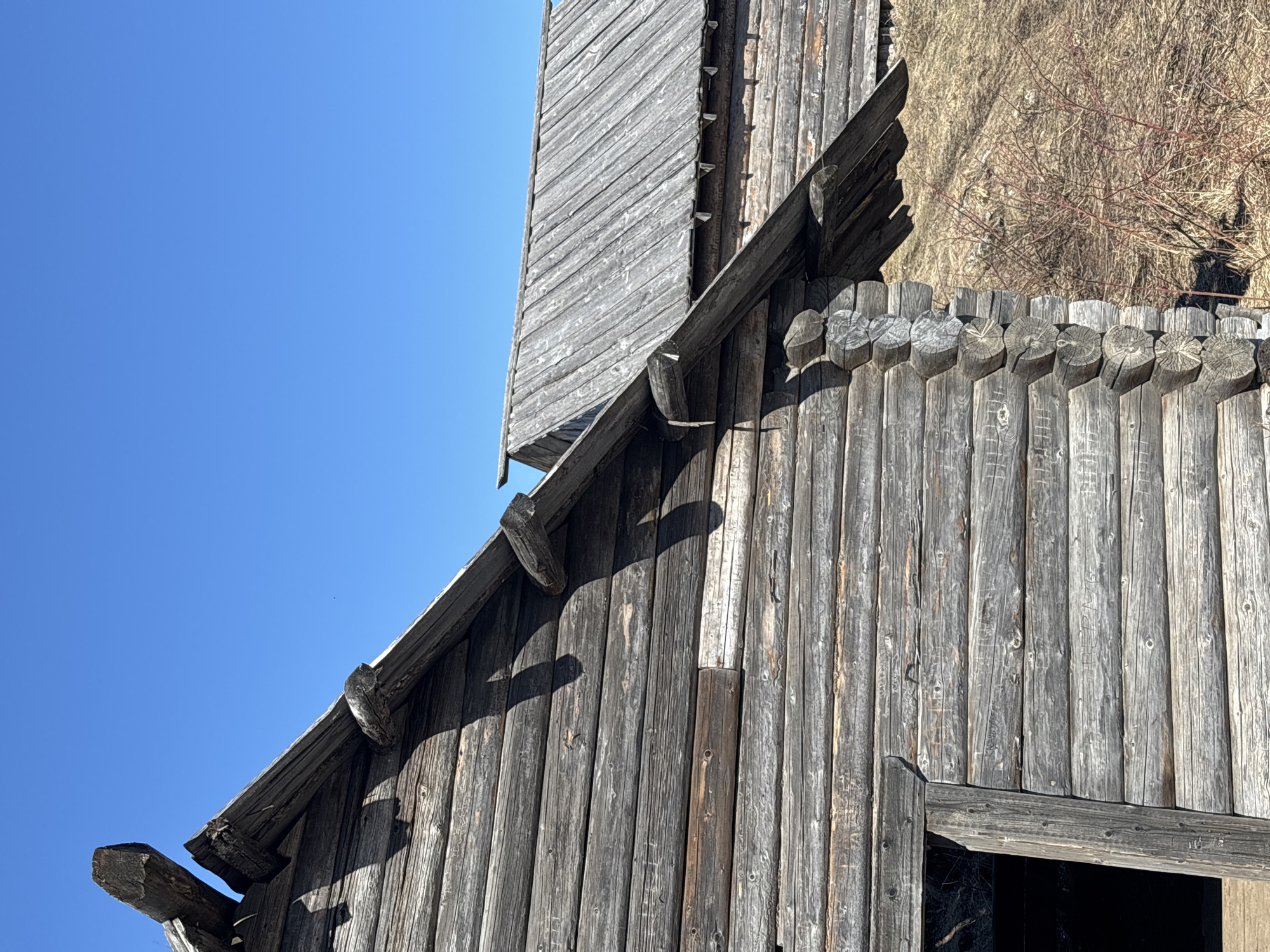 The image captures a weathered wooden structure in Gubakha, Russia, situated against a clear blue sky. The building's exterior is constructed from vertical logs or planks, exhibiting a gray, aged appearance. At the top of the frame, there is a section of horizontal logs, possibly a fence or part of the structure's design. Some logs have carved or inscribed surfaces. The structure features a sloping roof with what appears to be a drainage system. There's a glimpse of dry grass or vegetation behind the structure. The image is a mid-angle shot, focusing on the details of the wood and the building's design.