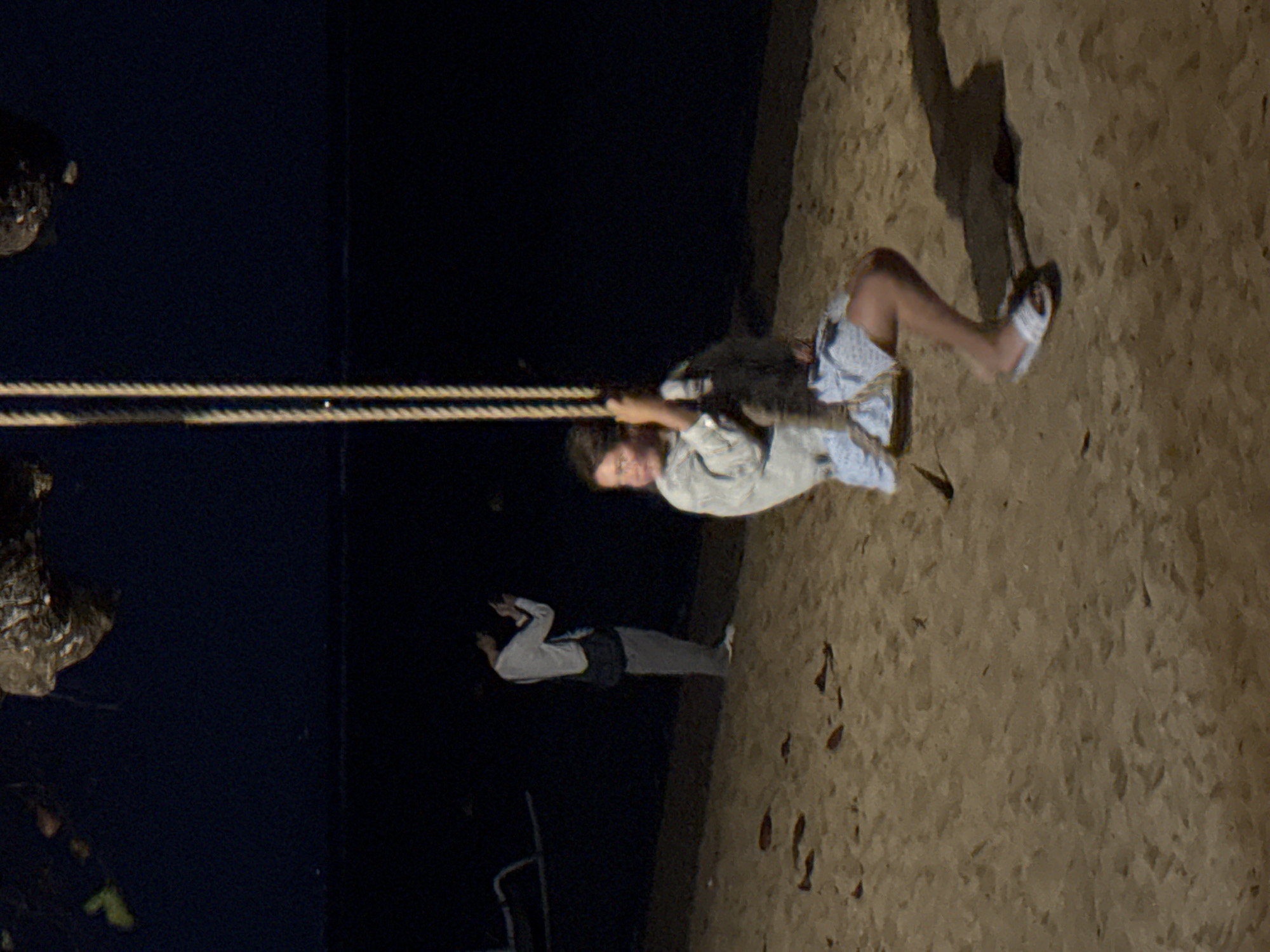 In Denpasar, Indonesia, on a beach at night, a young person is swinging on a rope swing. They are wearing a light-colored jacket and shorts with a blue and white pattern, and white slide sandals. Their left hand is gripping the rope, and their legs are bent. Another person stands in the background on the left, wearing a white long-sleeved shirt, grey pants, and a dark bag slung across their shoulder, holding a phone to take a picture. The beach is sandy, and the ocean is visible in the dark background.