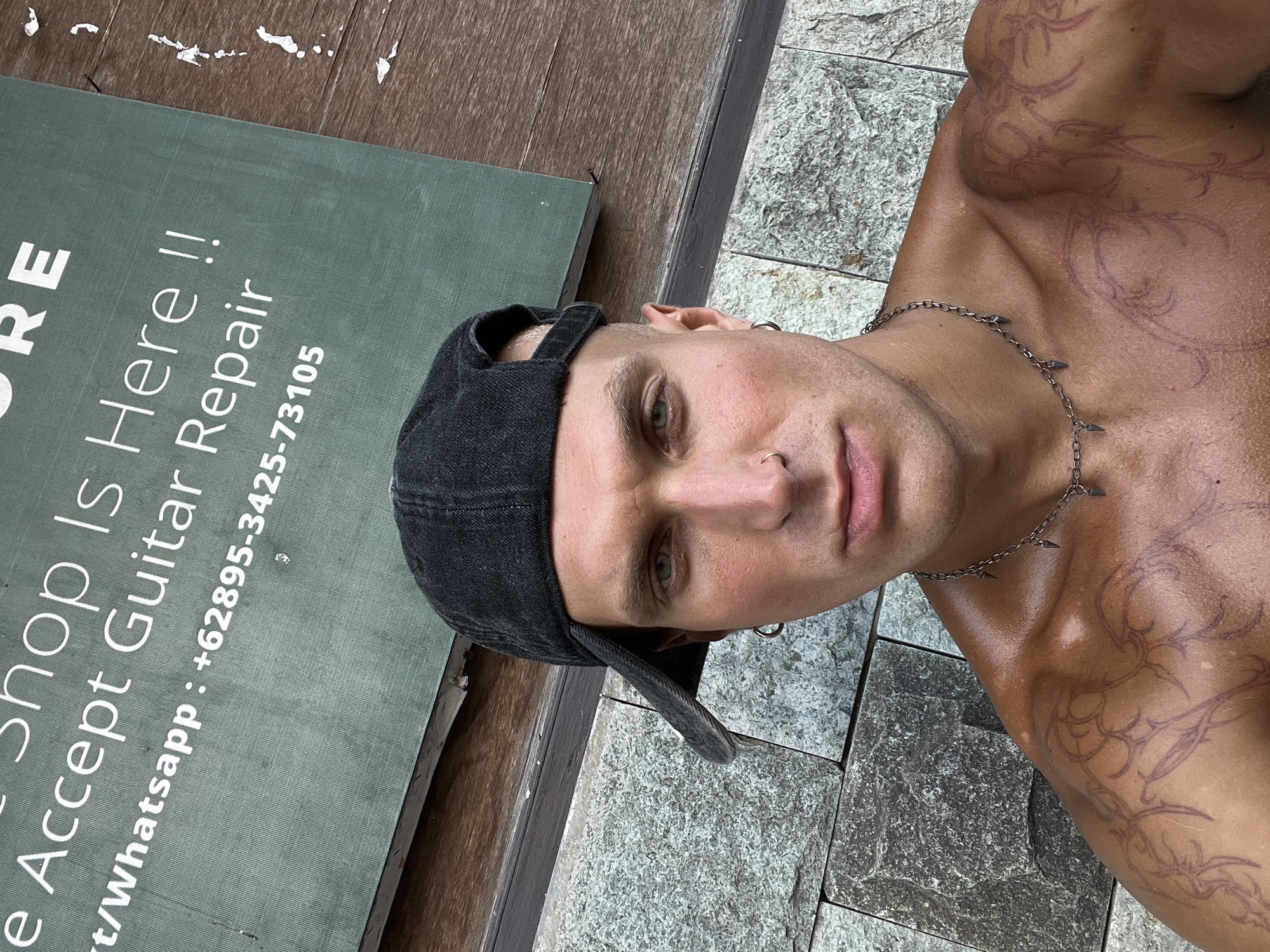 The image shows a man wearing a black baseball cap and a necklace, posing for a photo in front of a green sign. The sign has white text that reads, "Shop Is Here !! We Accept Guitar Repair t/whatsapp : +62895-3425-73105". The man has tattoos on his chest and shoulder. The background consists of a stone wall and a wooden surface. The man appears to be in an outdoor setting.