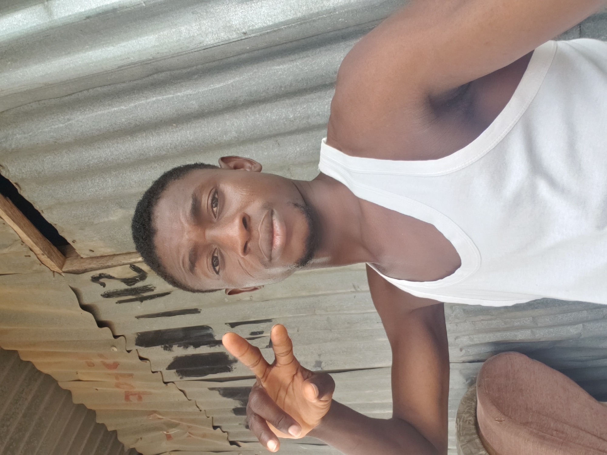 A man wearing a white tank top stands in what appears to be a modest dwelling with a corrugated metal roof. The man is making a peace sign with his hand. Writing is visible on the underside of the roof. The location is presumed to be Adankolo, Nigeria.