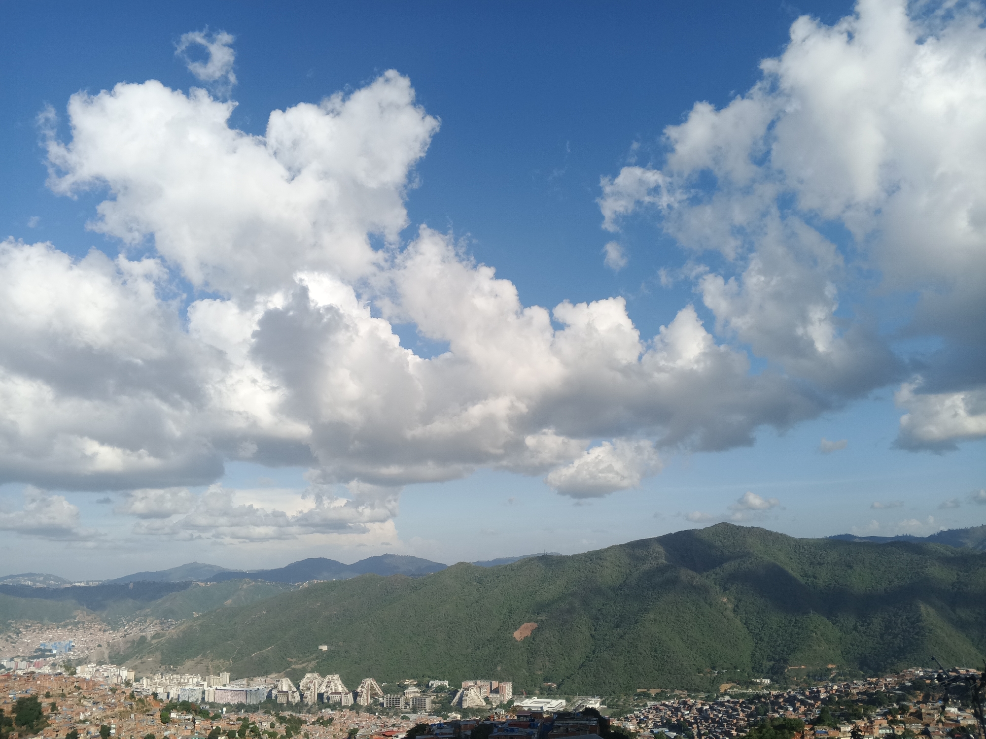 The image depicts an expansive view of Caracas, Venezuela, framed by a bright blue sky filled with fluffy, white clouds. Below the skyline, a series of green, mountainous ridges descend to meet the urban sprawl of the city. The foreground features a dense collection of buildings, many with red-tiled roofs, indicative of residential areas. In the midground, distinct architectural structures with a unique, pyramid-like design contrast with the surrounding buildings.