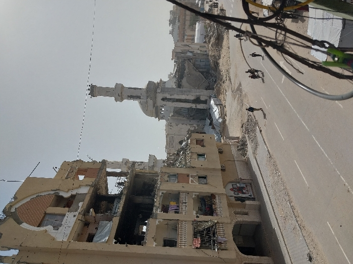 The image depicts a scene of extensive urban destruction in Khan Yunis, Palestinian Territory, under a clear, bright sky. In the foreground on the left, a multi-story building stands severely damaged, with large sections of its facade and internal structures exposed. Multiple floors show blast damage, shattered windows, and displaced debris, while a balcony on a lower floor displays hanging laundry items. Adjacent to this structure, a debris-strewn sidewalk borders a two-lane paved road, marked with white lines. Several individuals are walking on this road, moving away from the viewer: one person is on the left, a group of three to four people is near the center, and another lone individual is further down on the right. In the mid-ground, beyond the road, a significant portion of a mosque is visible, featuring a prominent minaret and dome, both showing heavy structural damage and partial collapse amidst surrounding rubble. Further into the background, additional damaged buildings and debris piles are evident. A cluster of dark wires and cables partially obscures the extreme right foreground.