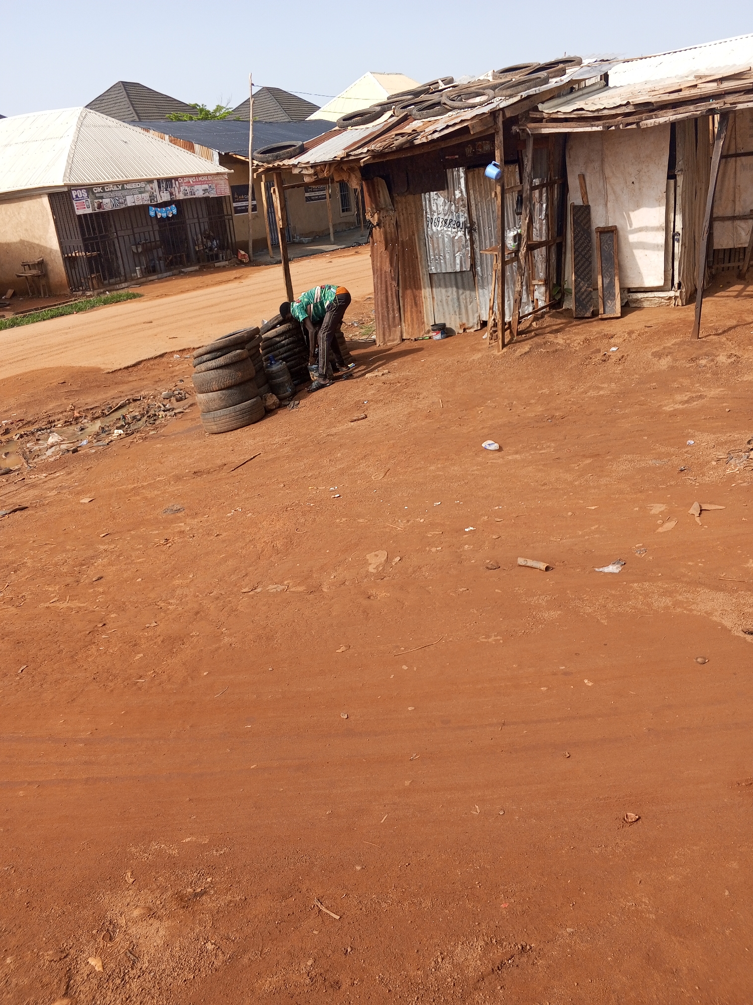 The image depicts a street scene in Abuja, Nigeria, characterized by red dirt roads and simple structures. In the foreground, a person wearing a green and black striped shirt and dark pants is bent over a stack of car tires. They appear to be working on or around the tires, though the specific action is not entirely clear. Behind the tires, several large blue plastic containers are visible.
To the left of the tires, a building with a white corrugated metal roof and beige walls is visible. It has barred windows and signage that reads "POS CK DAILY NEEDS" and "CK DRUGS & MORE ENTERPRISE". Several chairs and tables are set up outside this building. Further to the left, another building with a similar roof is present.
To the right of the tires, a makeshift structure constructed from corrugated metal sheets and wooden planks stands. Tires are also present on the roof of this structure, and a blue cup hangs from a hook on its side. Two wooden boards with writing on them are leaning against the structure. The ground throughout the scene is covered in red dirt, with some scattered debris and a small puddle of murky water to the left. The sky is a pale, clear blue, suggesting a bright, possibly warm, day.