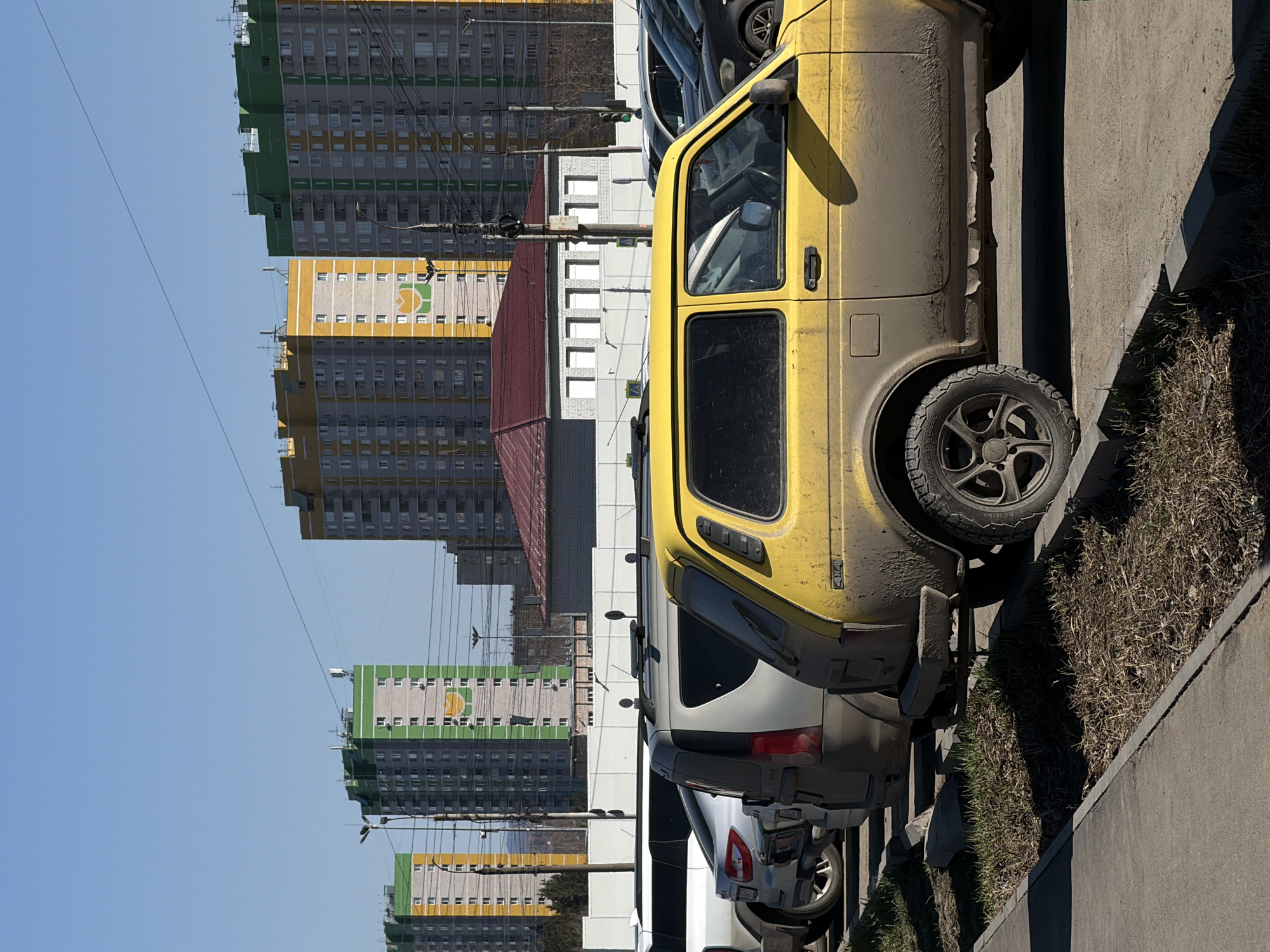 The image captures a street scene in Izhevsk, Russia, with several cars parked along a curb. Dominating the foreground is a dirty yellow compact car. Behind it are glimpses of other parked vehicles, including a silver SUV. In the background are several multi-story apartment buildings with a repeating pattern of green, yellow, and grey panels. Utility poles and wires are visible against the clear blue sky. The scene appears to be captured on a sunny day. The vehicles are parked along a concrete curb.