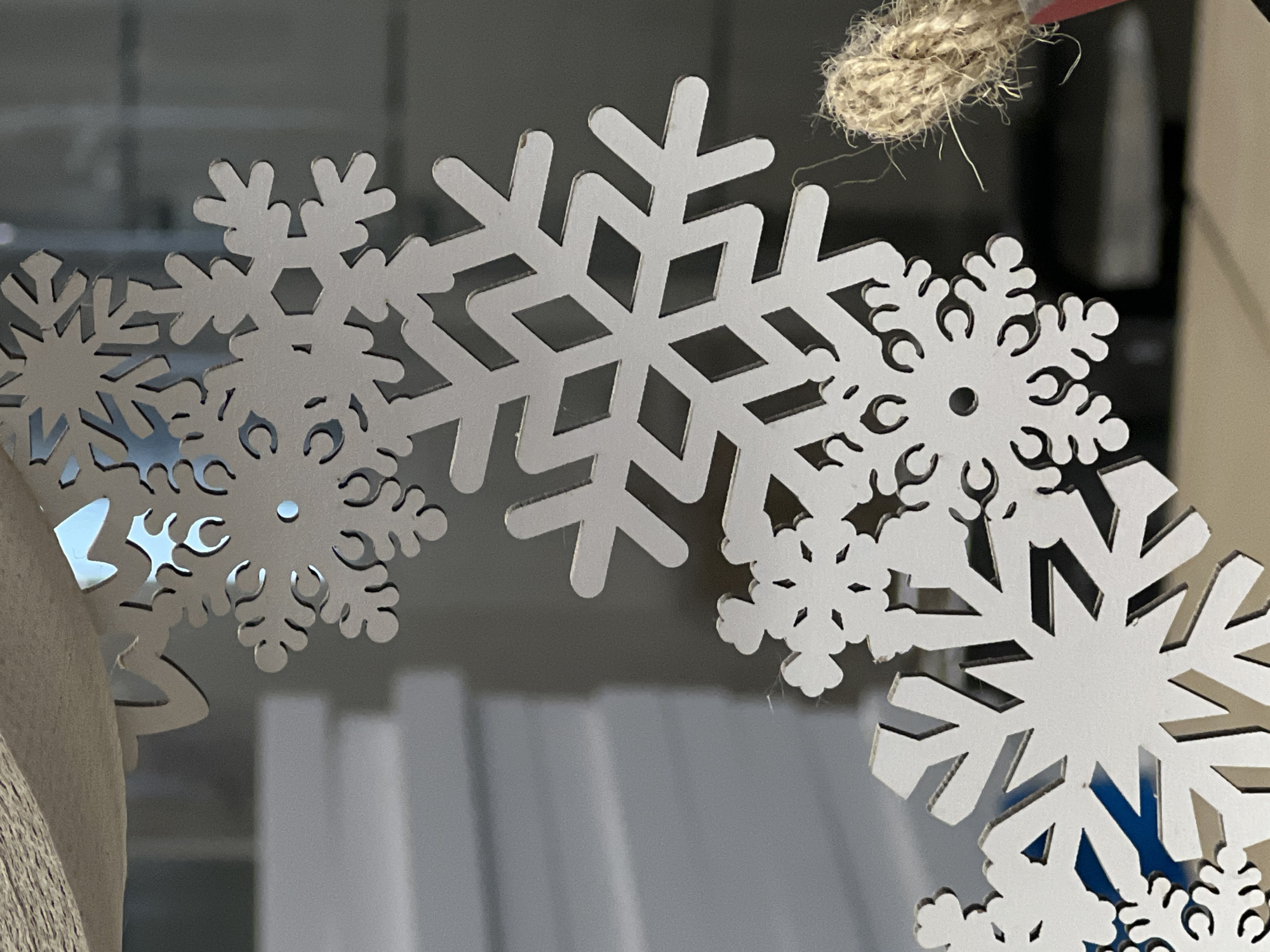 The image presents a close-up view of multiple white, intricate snowflake decorations, likely crafted from wood or plastic. These artificial snowflakes are designed with various detailed cut-out patterns, showcasing different hexagonal and branching arm structures, some featuring smaller, ornate details or central holes. They appear to be part of a larger decorative piece, possibly a wreath or garland, given their arrangement in an arc and some overlapping.
In the upper right corner, a segment of thick, braided jute twine or natural rope is visible, suggesting a hanging mechanism for the decoration. The background is softly blurred, creating a shallow depth of field that emphasizes the sharpness and detail of the snowflakes. The indistinct background shows muted shades of grey and white, with vague vertical forms that could indicate window blinds, reflections, or other architectural elements in an indoor setting. The lighting is even, illuminating the textures and details of the snowflakes without harsh shadows. No people or visible text are present. The overall scene evokes a festive, winter holiday theme.