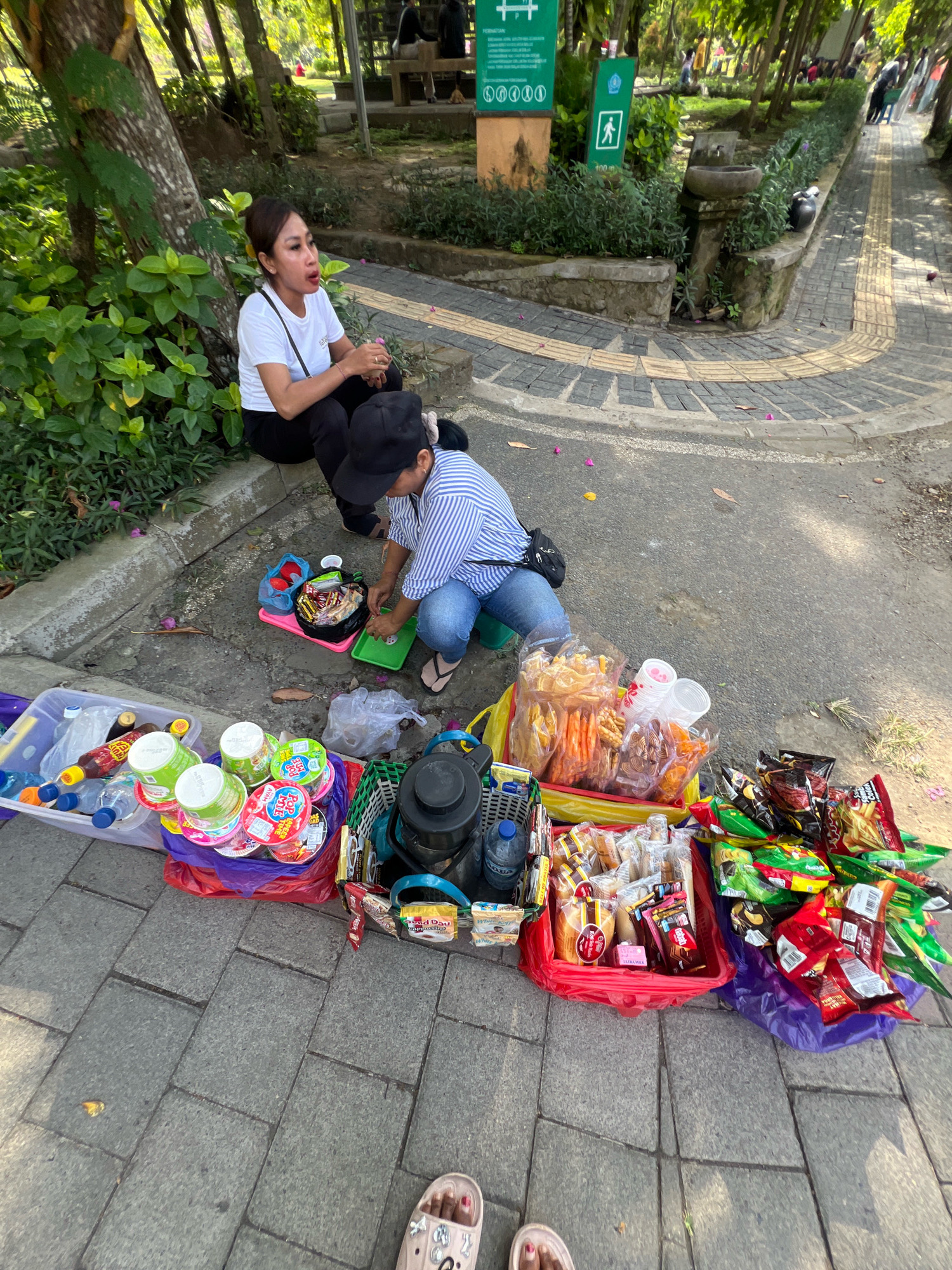 Two women are sitting on the side of a paved walkway in a park-like setting, likely in Denpasar, Indonesia, selling snacks and drinks. The woman on the left is wearing a white t-shirt and black pants, and is looking to her right. The woman on the right is wearing a blue and white striped shirt and jeans, and is focused on preparing items on a small green tray. A variety of packaged snacks, drinks in bottles and cups, and other items are laid out on the ground in front of them, suggesting a small roadside business. The ground is paved with large grey rectangular stones, and there is a curb with green foliage. In the background, trees and more park benches are visible, with other people in the distance. The lighting indicates it is daytime, with sunlight filtering through the trees.