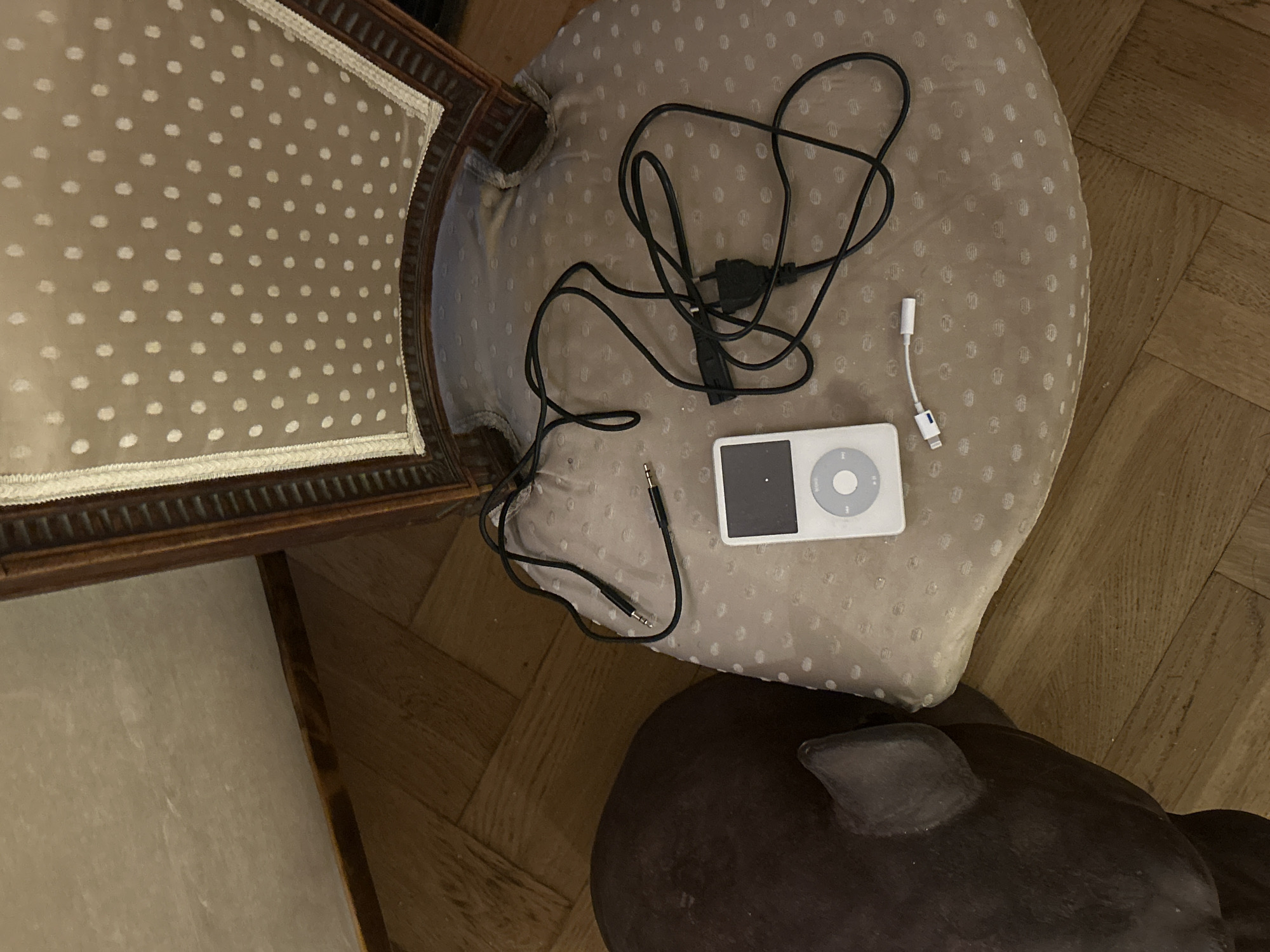 An overhead shot depicts a partially visible ornate chair with a patterned seat cushion, upon which an iPod is placed alongside various charging cables and a white dongle. The setting appears to be indoors, likely a domestic interior, with a parquet floor visible in the background. A dark, curved object of indeterminate nature occupies the lower right corner. The location is Unknown, Unknown.