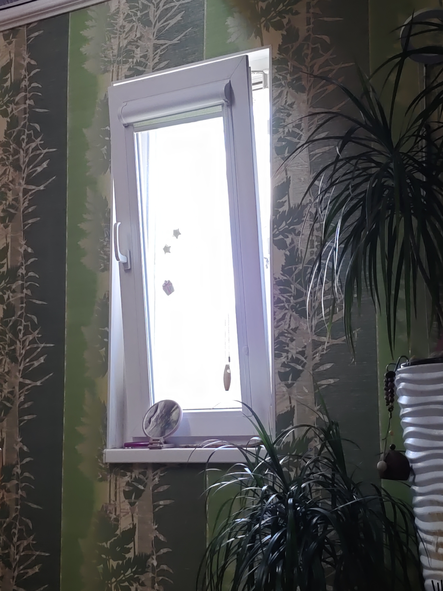 The image shows a partially open window in a room with striped wallpaper. The window frame is white, and the windowpane is open at an angle, allowing bright light to enter. On the windowsill, there is a small round mirror with a pink stand. To the right of the window, a large potted plant with long, dark green leaves is visible. Behind the plant, a textured white vase can be seen. The wallpaper features a pattern of stylized trees and leaves in varying shades of green. Several small decorative items, including what appear to be stars and a gift box, are affixed to the inside of the windowpane. A string with beads and a small wooden ornament hangs from the top right corner of the window frame. The overall scene suggests an indoor setting, possibly a domestic environment. The image does not provide specific visual cues to pinpoint its exact location within Rostov-on-Don, Russia.
