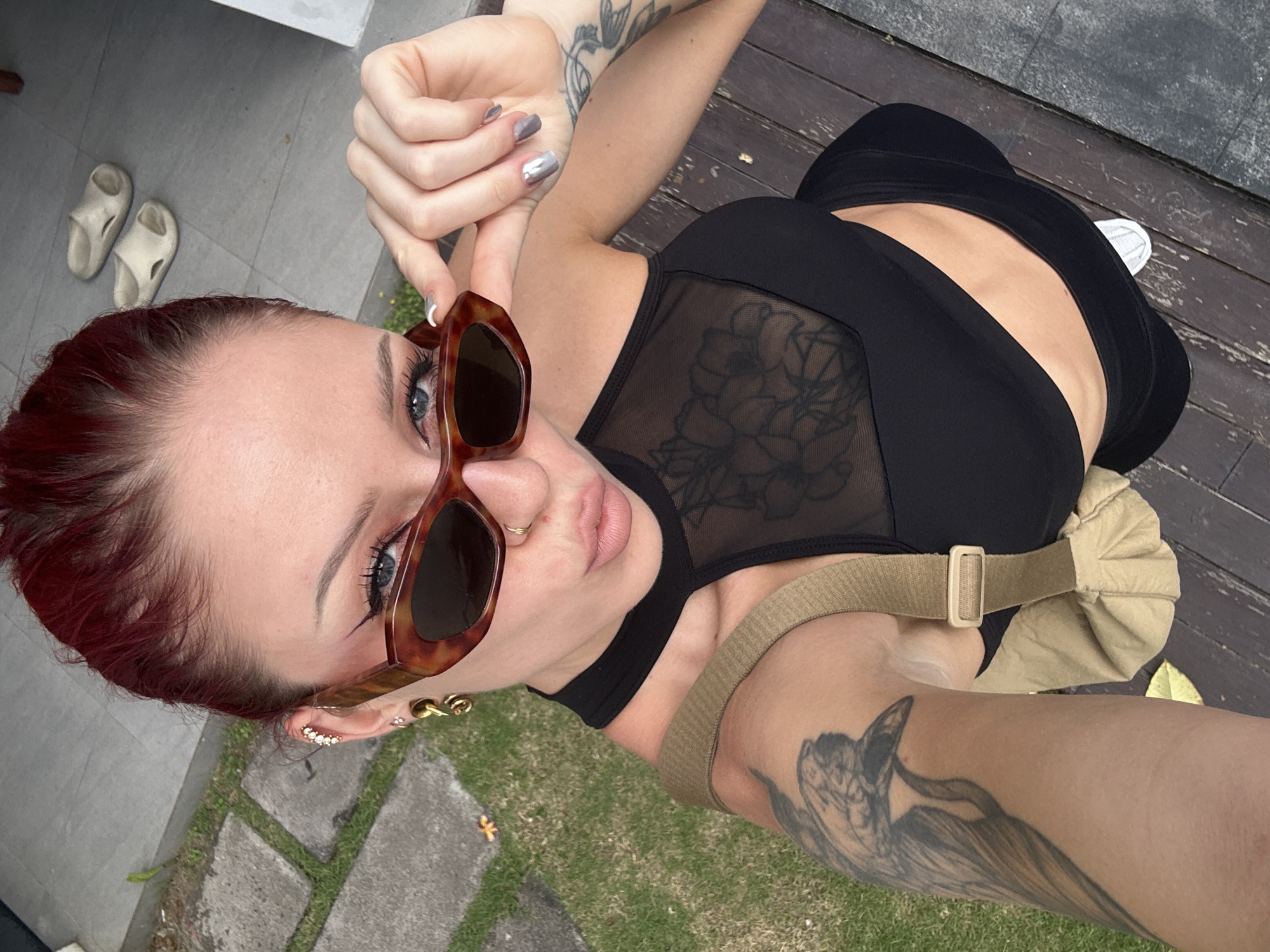A woman poses for a selfie in North Kuta, Indonesia. She is wearing a black crop top with a mesh floral design, black pants, and brown sunglasses. She has red hair, a nose ring, and tattoos on her arms. Her fingernails are painted silver. She is wearing multiple earrings on her left ear, and is wearing a tan-colored crossbody bag. She is standing on a wooden deck next to a grassy area and a concrete walkway where a pair of white slip-on shoes are visible.