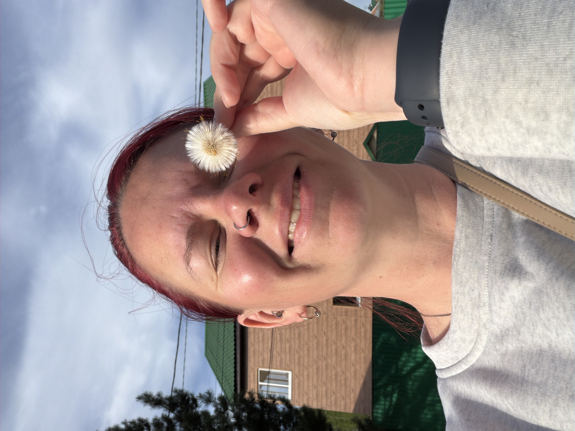 A woman is holding a dandelion clock up to her face. She has red hair, fair skin, and a nose piercing. She's wearing a grey sweatshirt and a black fitness tracker on her wrist. The background includes a wooden building with a green roof and a blue sky with clouds. The specific location is unknown and cannot be determined from the image.