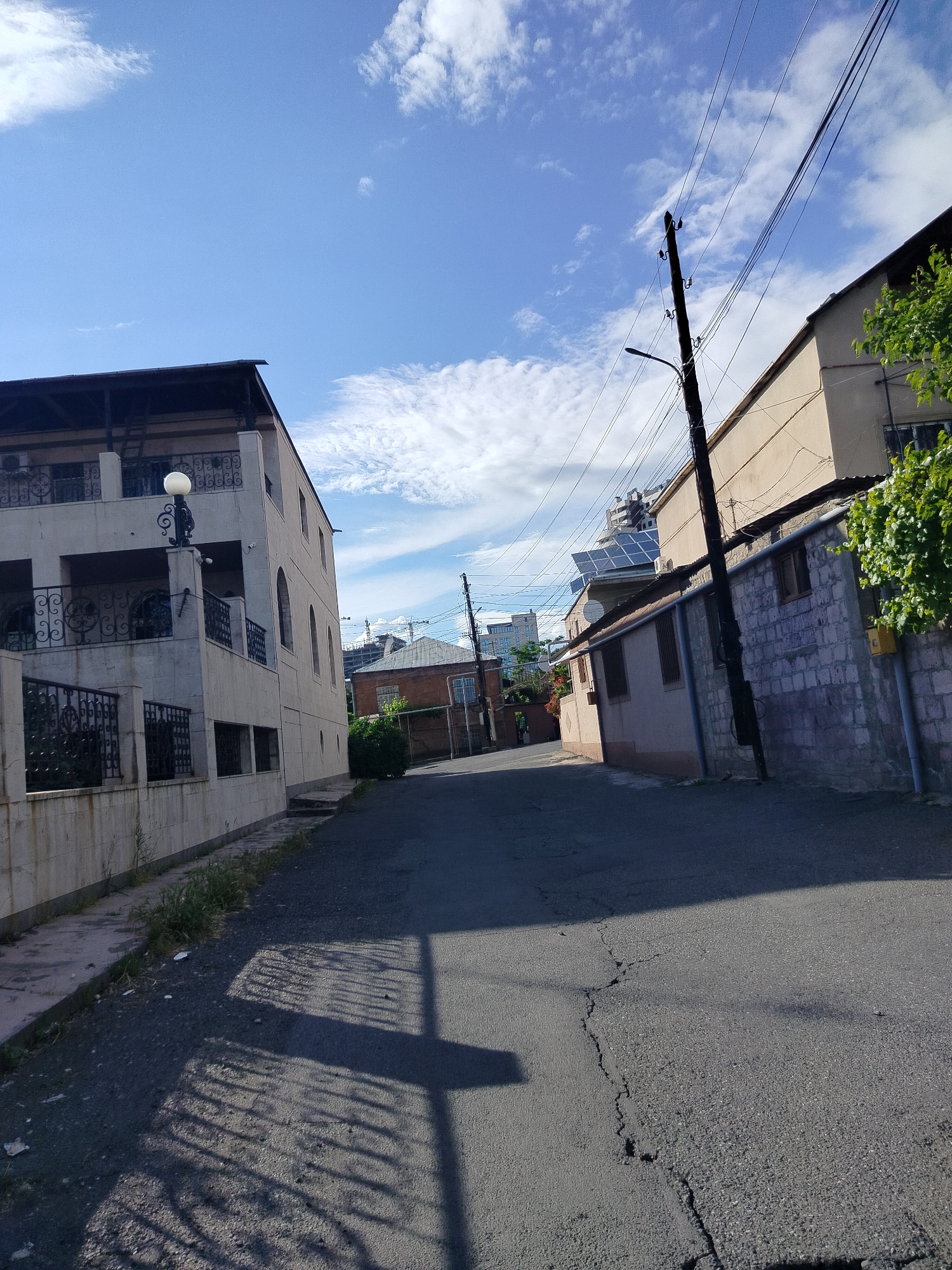 The image depicts a street in Yerevan, Armenia, on a sunny day. The street is asphalt, with cracks in the pavement, and slopes gently downward. A two-story building with a decorative wrought-iron balcony is on the left. On the right is a single-story structure with a brick and stucco facade. Power lines run along the street, supported by wooden poles. In the distance are taller buildings and construction cranes. There are no people visible in the immediate foreground of the street. Shadows of trees and poles are cast on the road.