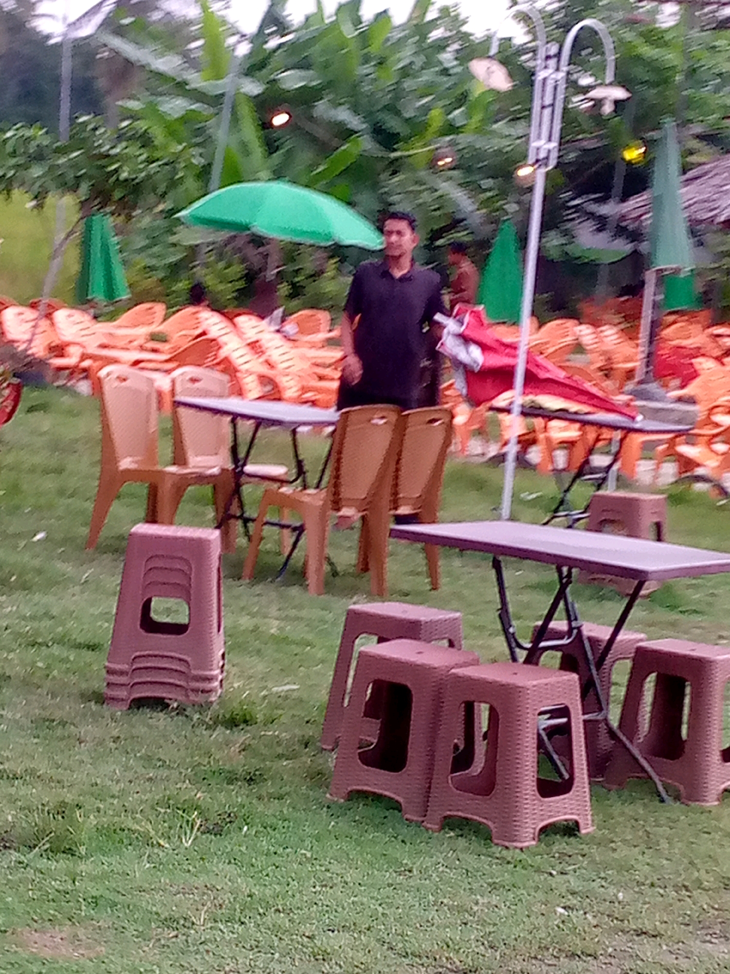 The image depicts an outdoor seating area, likely a restaurant or cafe, with a grassy ground. In the foreground, several brown plastic stools are arranged, some stacked. A long, dark table with a metal folding base is positioned behind the stools. A man in a dark t-shirt is standing near the table, and behind him, there are rows of stacked orange plastic chairs. Green umbrellas are visible in the background, along with lush green foliage from trees. A street lamp with multiple lights is also present. The scene suggests a relaxed, possibly casual, dining or social setting.