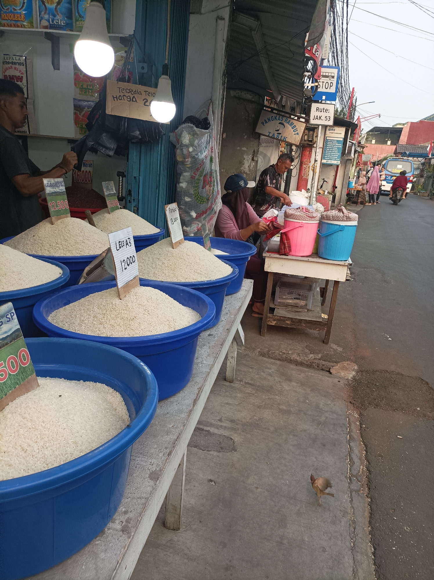 A street-level view of a small market stall in West Jakarta, Indonesia, shows piles of rice in blue plastic tubs displayed on a wooden counter. A man in a dark t-shirt is seen to the left, appearing to be working at the stall. Behind him, a blue metal shutter is visible. Two people are seated at a table to the right, with pink and blue cooler boxes. One person, wearing a pink headscarf, is handling what appears to be cloth or plastic. The scene is set along a moderately busy street with vehicles and pedestrians in the background. Overhead, a tangled network of electrical wires is prominent. A bus stop sign with "STOP" and "Rute: JAK. 53" is attached to a pole. A small bird is walking on the ground in the foreground.