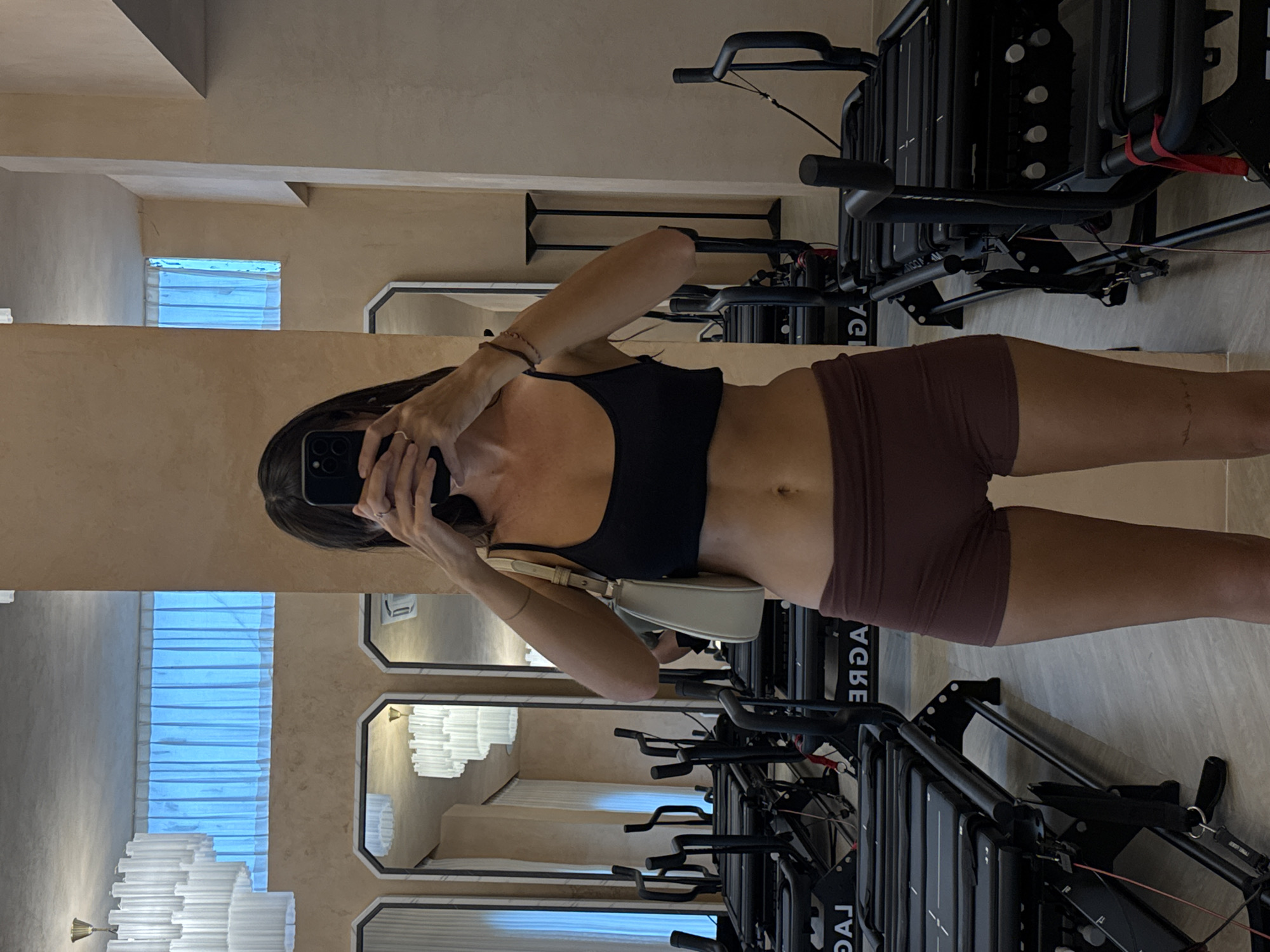 A tanned-skinned woman with long dark hair, wearing a black sports bra and dark purple shorts, holds a phone to take a photo of herself in a mirror. She holds a cream-colored purse at her side. She stands in a gym with multiple black Lagree fitness machines. The background shows a mirrored wall with reflections of a window with horizontal blinds and a tiered chandelier. The gym is likely located in North Kuta, Indonesia.