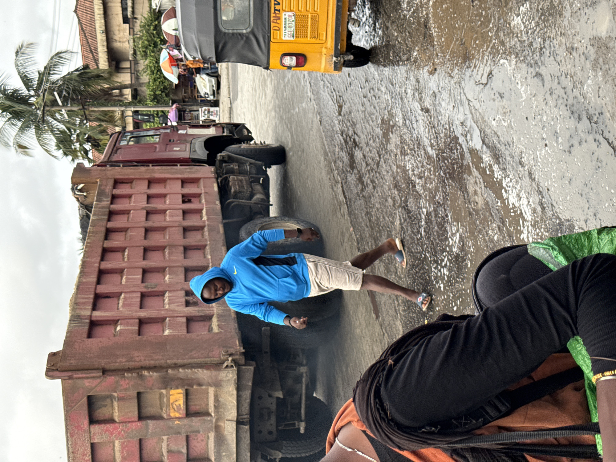 A partially rotated medium shot captures a street scene in Ado, Nigeria, featuring two individuals, a dump truck, and a three-wheeled taxi. One individual wearing a bright blue hooded sweatshirt and tan shorts stands between the first axle wheel of the dump truck with the bed raised to the right, with their legs spread and arms at their sides. Their gaze is directed towards the camera. The second individual is seated, seen from the shoulders down. They have dark hair under a black helmet. The dump truck is red, with a raised bed. The ground appears to be wet and reflective. In the background, a yellow three-wheeled taxi is partially visible, along with buildings and a palm tree.