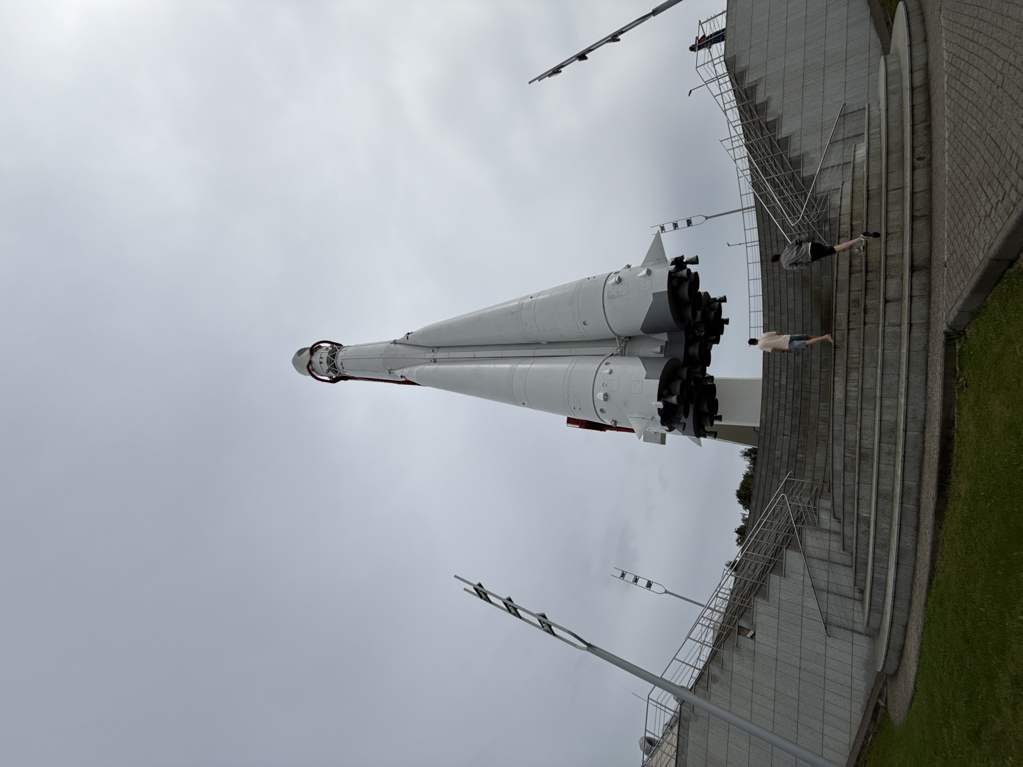 A low-angle, full shot captures a large, white rocket monument with red accents standing tall against a cloudy, overcast sky. The rocket is positioned on a raised platform, accessible by a wide, curved staircase. Two young men are ascending the stairs from the foreground, one slightly ahead of the other. A third person, appearing as a distant figure, stands on the upper platform near the rocket. The staircase is flanked by modern-looking railings and a gray tiled wall. To the left and right of the staircase, tall, minimalist lamp posts with multiple lights are visible. The foreground features a strip of green grass bordering a paved walkway. This scene is located at the State Museum of the History of Cosmonautics in Kaluga, Russia.