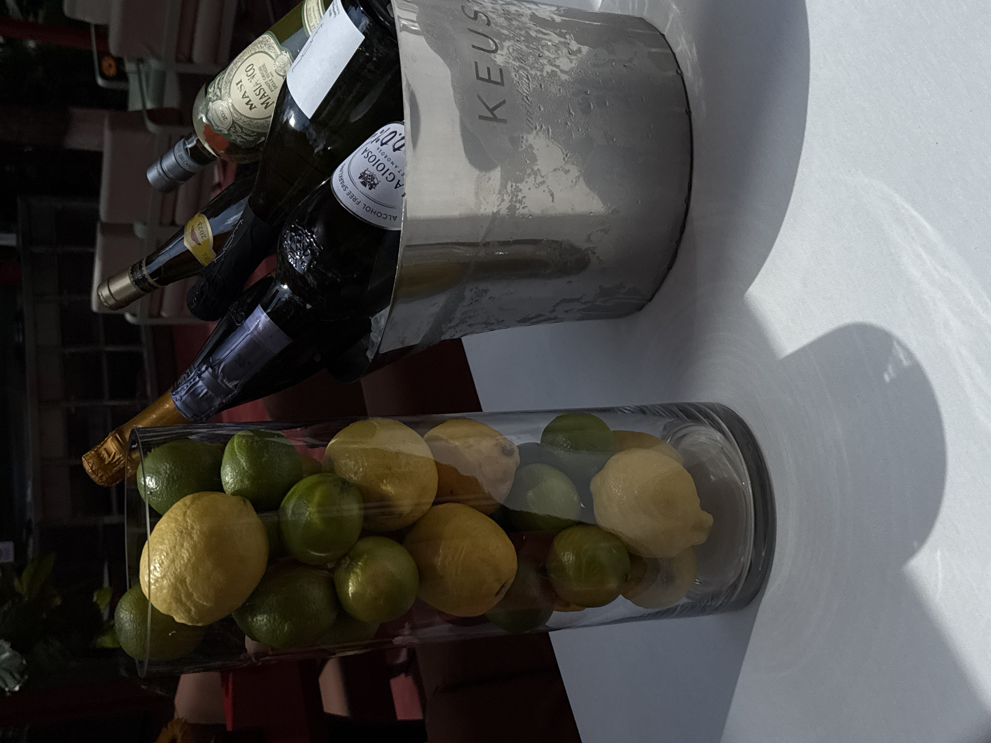 The image shows a scene on a white surface, likely a table. There's a silver ice bucket containing bottles of wine, including one labeled "MASI MASIANCO" and another marked as "ALCOHOL FREE SPARKLING...AGIOIOSA." The bucket has condensation droplets on its surface. In front of the bucket is a clear glass vase filled with lemons and limes. The setting seems to be in a dim area with some light reflecting on the white surface. This image does not contain information that confirms the location being in Yerevan, Armenia.