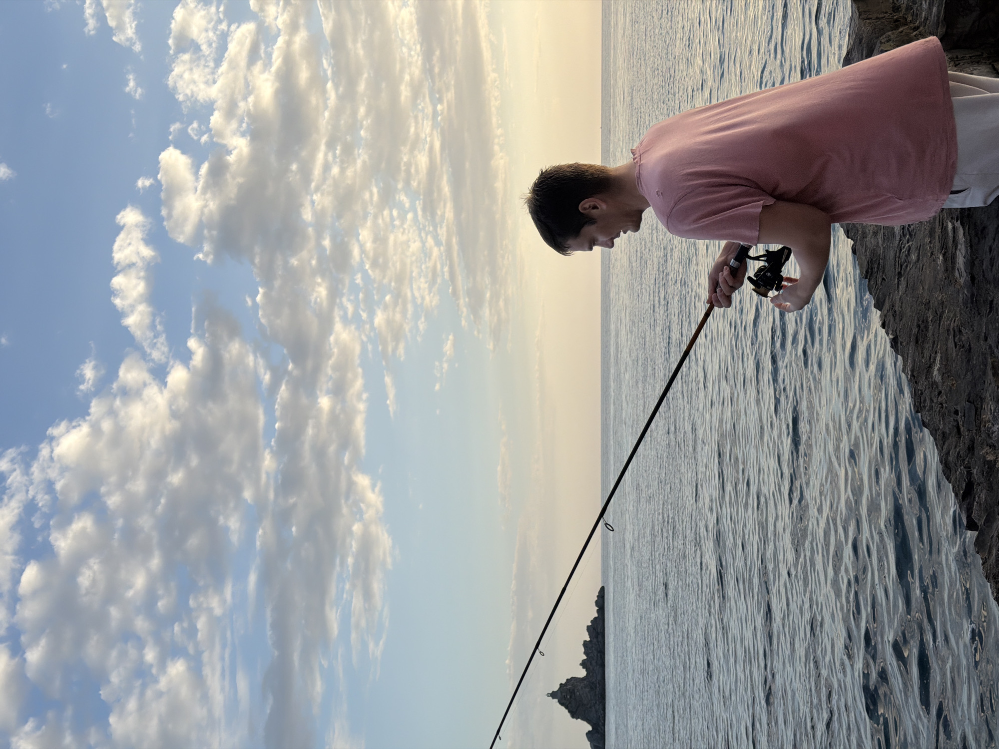 A man is fishing in Petrovac centar, Montenegro. He is standing on a rocky shoreline, holding a fishing rod and reel. The water is calm, and the sky is filled with clouds.