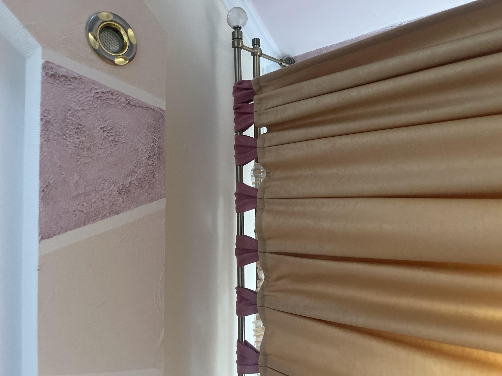 The image shows a close-up of a ceiling with textured pink and white geometric panels. A recessed ceiling light fixture with a gold rim is visible on the right. Below the ceiling, a metal curtain rod is mounted, from which hangs a curtain. The curtain is a pale beige color and has pleats. The top of the curtain has loop tabs in a darker pink color. The scene appears to be an interior of a room. There are no people or actions visible. The location of Tarnivtsi, Ukraine is not depicted in the image.