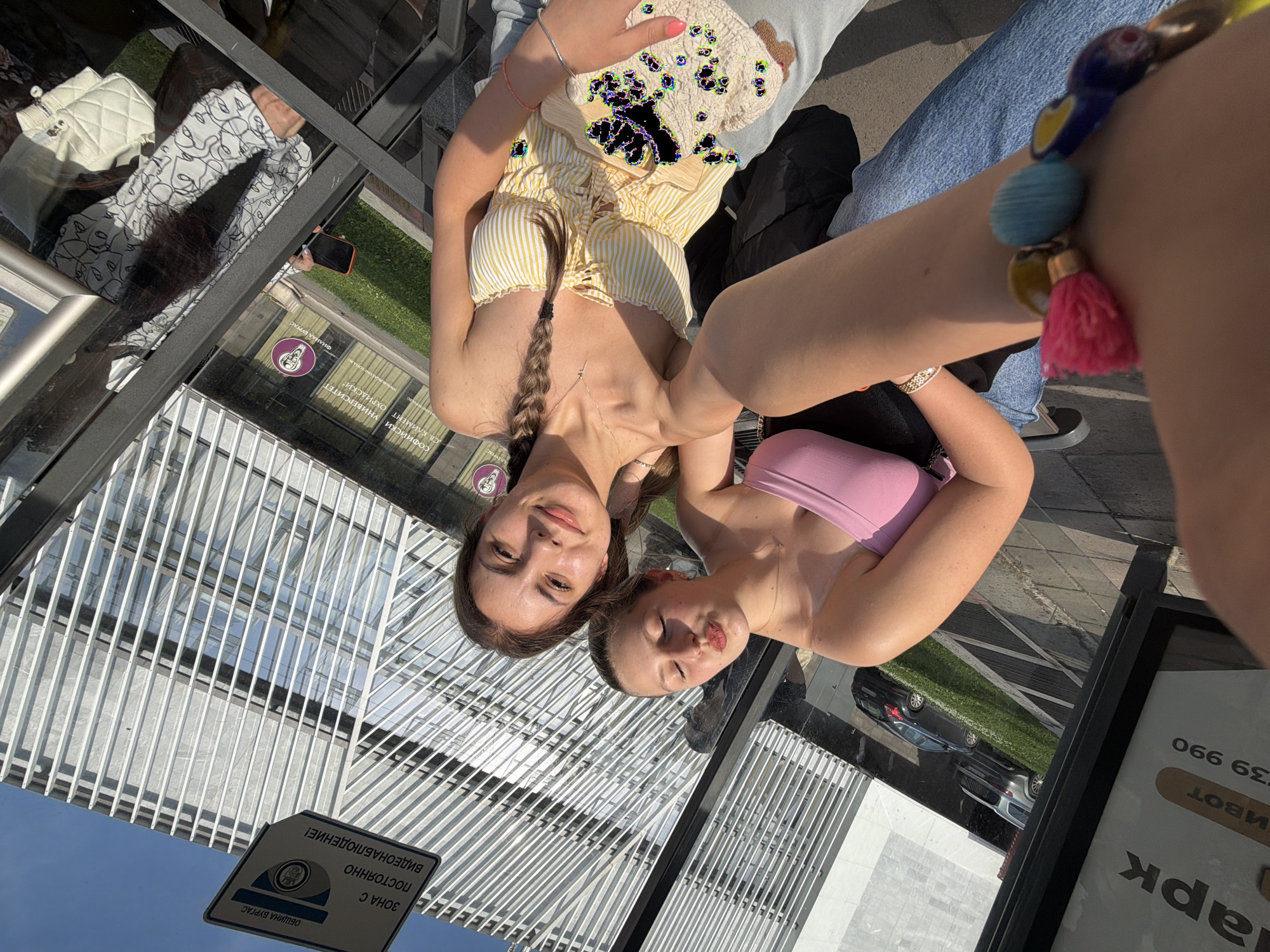 The image, captured from an elevated perspective and rotated counter-clockwise, features two young women posing outdoors on a bright, sunny day. They appear relaxed and are looking towards the camera, possibly for a selfie.
The woman on the left has long dark hair, styled with a braid resting over her left shoulder. She wears a yellow and white striped, ruffled crop top with a laced front and a delicate necklace. A simple bracelet is visible on her right wrist, and she holds a light-colored, textured item, possibly a small bag or decorative cushion.
The woman on the right wears a pink strapless top. Her right arm, adorned with a vibrant, multi-colored beaded bracelet featuring several tassels (including a prominent pink one), is extended towards the foreground. Her face is slightly turned, displaying a playful pouting expression.
The setting appears to be an area with large reflective surfaces, such as a glass-enclosed terrace, building facade, or a modern bus stop shelter. These surfaces show clear reflections of the surroundings, including architectural structures with a grid pattern, a bright blue sky, green grass, and several cars.
Visible text from reflections and signs confirms the location context. Signs read "ОБЩИНА БУРГАС" (Municipality of Burgas) and "ЗОНА С ПОСТОЯННО ВИДЕОНАБЛЮДЕНИЕ!" (Zone with permanent video surveillance!). Additionally, "СОФИЙСКИ УНИВЕРСИТЕТ 'СВ. КЛИМЕНТ ОХРИДСКИ'" (Sofia University "St. Kliment Ohridski") and "ФИЛИАЛ БУ...", likely denoting a Burgas branch, are visible. Other partial text includes "арк", "Ивот", and "39 990". The bright lighting indicates it is daytime.