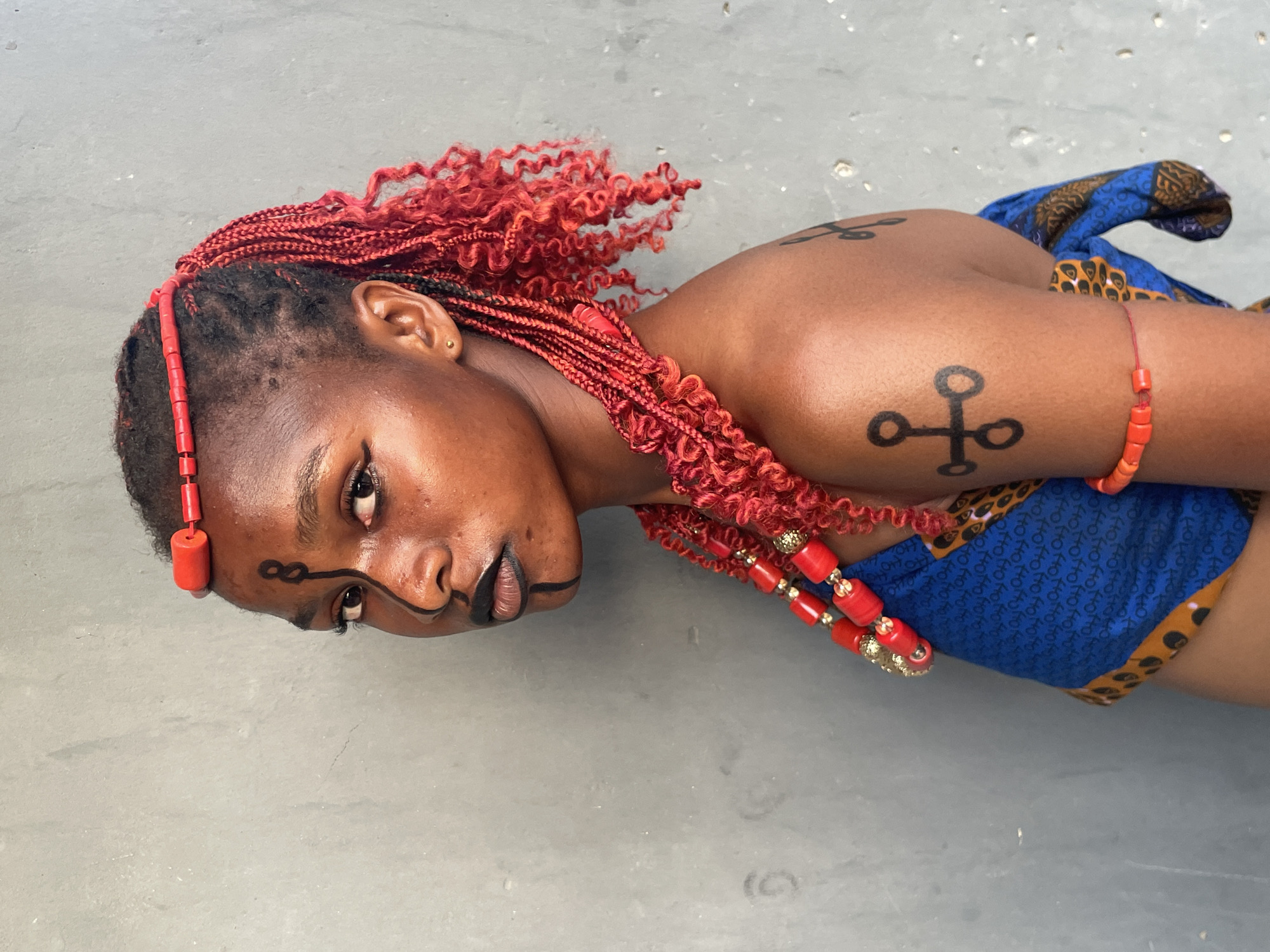 A close-up portrait features a young individual, likely female, with dark skin, positioned against a plain, light grey background, possibly a concrete surface. The subject is adorned with vibrant red beaded jewelry, including a headband or hair accessory, a long layered necklace with gold accents, and a bracelet. Their hair is styled in striking red braids or extensions, some curly, framing the face.
The individual's face is accented with black body paint or makeup, featuring thin lines extending from below the eyes, across the cheeks, and on the chin. Their gaze is direct and serious, looking towards the viewer. Two distinct black, circular, cross-like symbols are painted on their right arm, resembling traditional African motifs. The subject is partially clad in a vibrant blue fabric with a repetitive pattern, possibly a wax print, with hints of yellow or brown patterned fabric also visible. The overall aesthetic suggests a cultural representation or photoshoot, highlighting traditional adornment and body art from Badagry, Nigeria. The lighting is natural and even, emphasizing the details of the subject's features and accessories. No visible text is present in the image.
