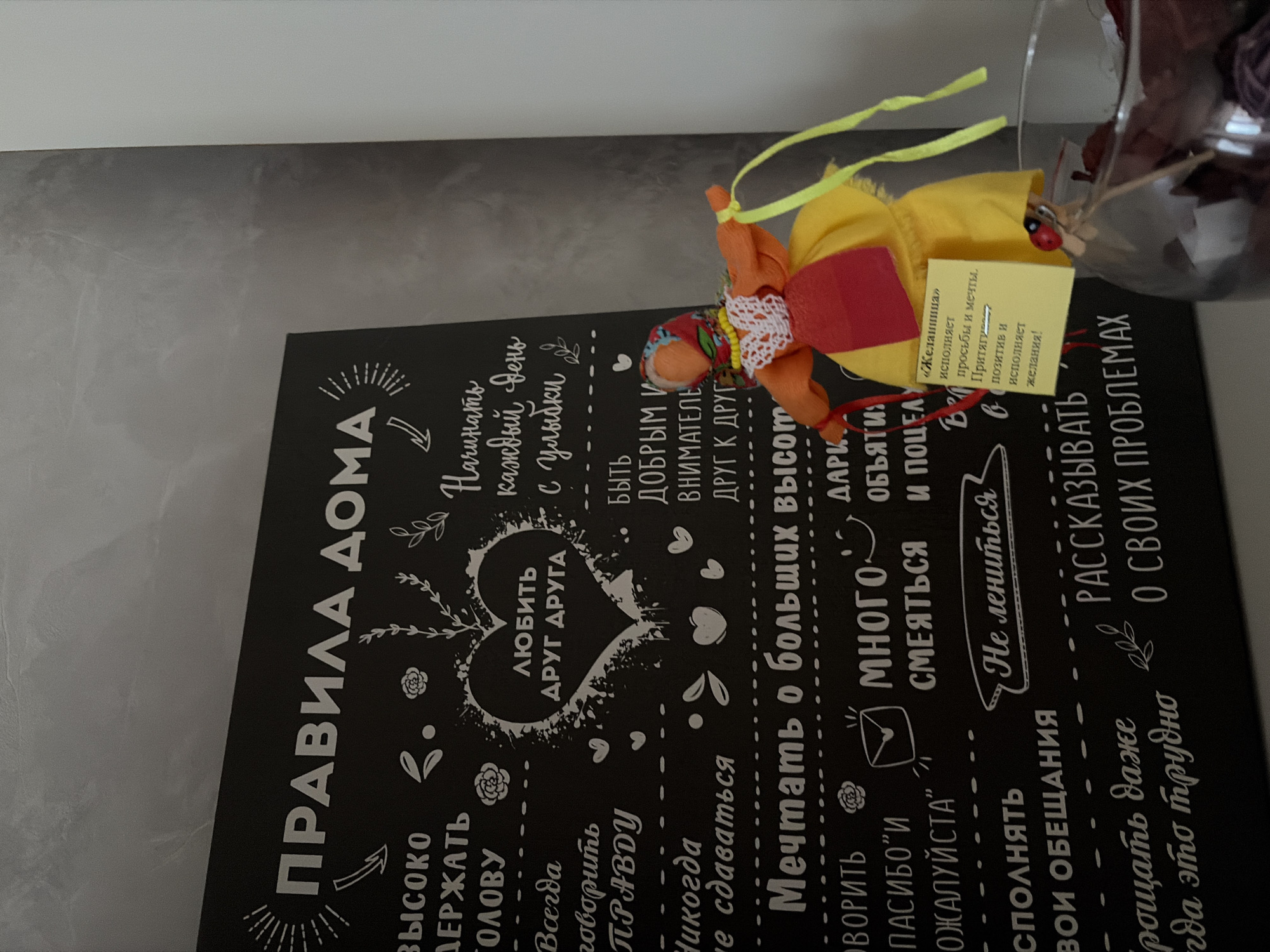 The image displays a close-up, slightly tilted view of a decorative black board with white lettering, likely a "House Rules" sign, positioned against a light gray background. On the right side, a vibrant, handcrafted fabric doll, appearing to be a traditional Slavic "wish doll" (желаниица), is partially visible. The doll is dressed in yellow, red, and orange fabrics, adorned with white lace and beads, and has yellow ribbons extending from its head. A small yellow tag is attached to the doll, featuring a tiny red ladybug. Behind the doll, a clear glass container, possibly holding dried plant material or potpourri, is partially seen.
The board's text is in Russian, titled "ПРАВИЛА ДОМА" (House Rules). The rules listed include: "ВЫСОКО ДЕРЖАТЬ ГОЛОВУ" (Keep your head high), "Всегда говорить ПРАВДУ" (Always tell the truth), "Никогда не сдаваться" (Never give up), "ЛЮБИТЬ ДРУГ ДРУГА" (Love each other, within a heart graphic), "Начинать каждый день с улыбки" (Start each day with a smile), "БЫТЬ ДОБРЫМ И ВНИМАТЕЛЬНЫМ ДРУГ К ДРУГУ" (Be kind and attentive to each other), "Мечтать о больших высотах" (Dream big), "ГОВОРИТЬ "СПАСИБО" И "ПОЖАЛУЙСТА"" (Say "Thank you" and "Please"), "МНОГО СМЕЯТЬСЯ" (Laugh a lot), "ДАРИТЬ ОБЪЯТИЯ И ПОЦЕЛУИ" (Give hugs and kisses), "Не лениться" (Don't be lazy), "ИСПОЛНЯТЬ СВОИ ОБЕЩАНИЯ" (Keep your promises), "ПРОЩАТЬ ДАЖЕ КОГДА ЭТО ТРУДНО" (Forgive even when it's hard), and "РАССКАЗЫВАТЬ О СВОИХ ПРОБЛЕМАХ" (Talk about your problems).
The tag on the doll reads: "«Желаниица» исполняет просьбы и мечты. Притягивает позитив и исполняет желания!" (translation: "«Wish Doll» grants wishes and dreams. Attracts positivity and grants wishes!"). The scene suggests an indoor domestic setting, possibly a decorative corner in a home in Perm, Russia, given the cultural context of the doll and the Russian text.