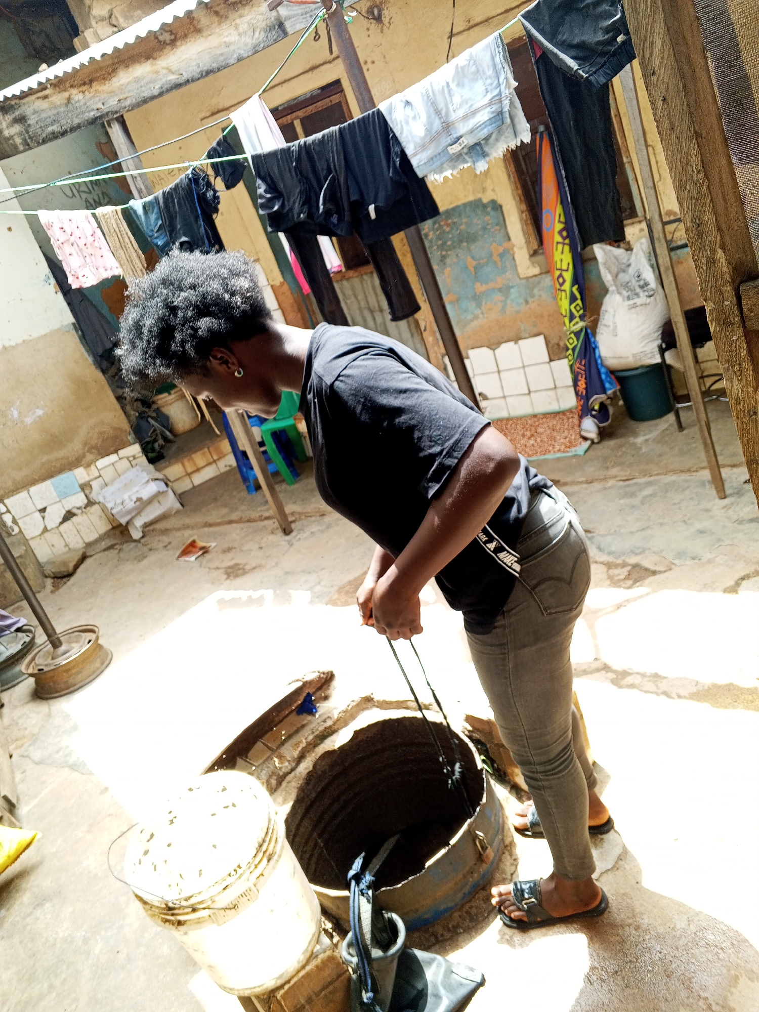 The image depicts a woman standing outside next to a well in Gwong, Nigeria. She is wearing a black t-shirt, grey jeans, and sandals, and is leaning over the well, holding a rope or cable. The well is made of a metal barrel, and there are several other objects nearby, including a white bucket, various items hanging on a clothesline above, and a blue plastic chair. The courtyard area is concrete, and the background shows weathered buildings.