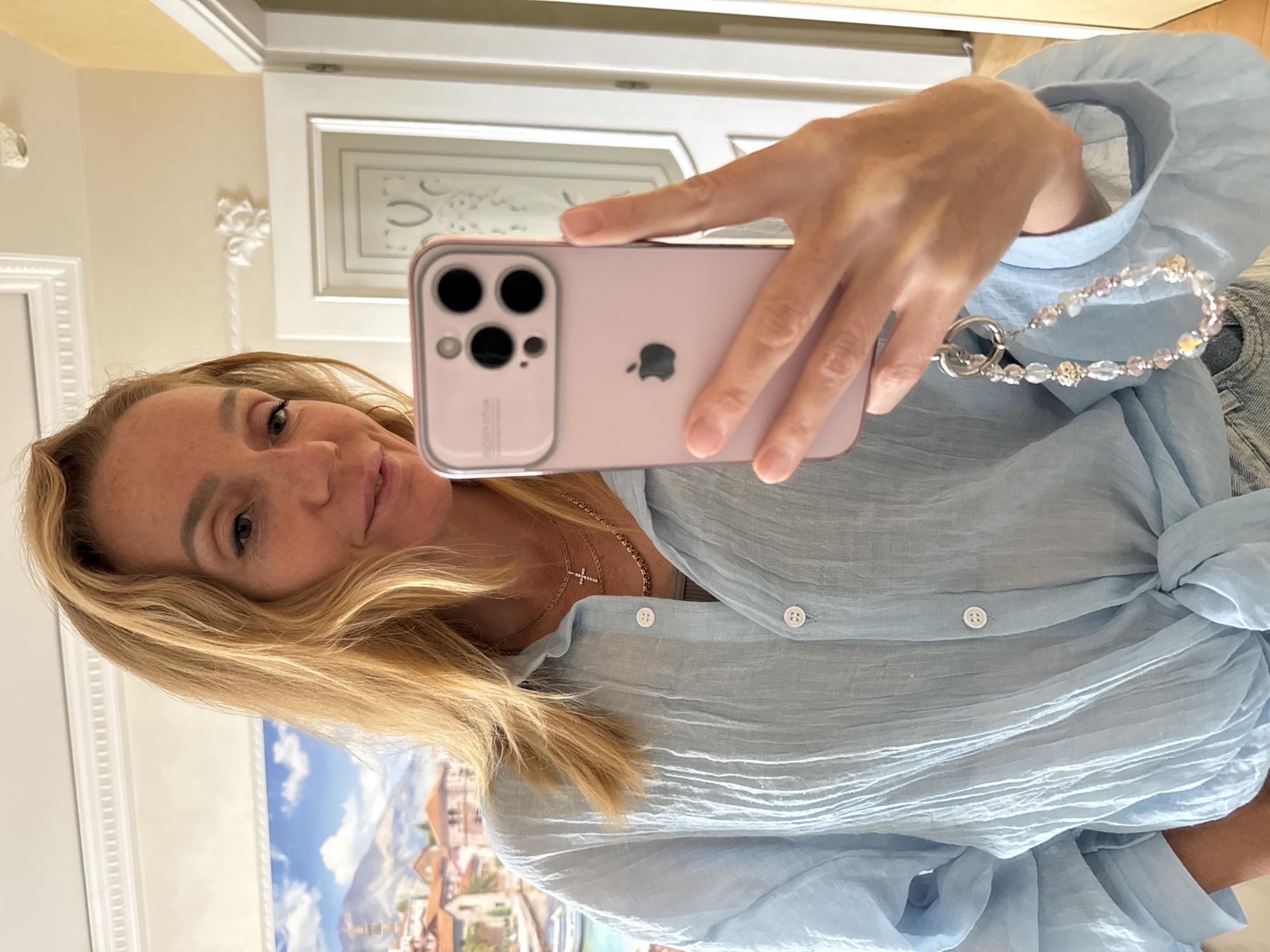 An unseen photographer takes a mirror selfie holding an iPhone with a pink case and a beaded charm. The person in the photo is a woman with blonde hair, wearing a light blue crinkled button-up shirt tied at the waist and two necklaces, one with a cross pendant. She is indoors, with a painting of a European town on the wall behind her, and a doorway visible to her right.