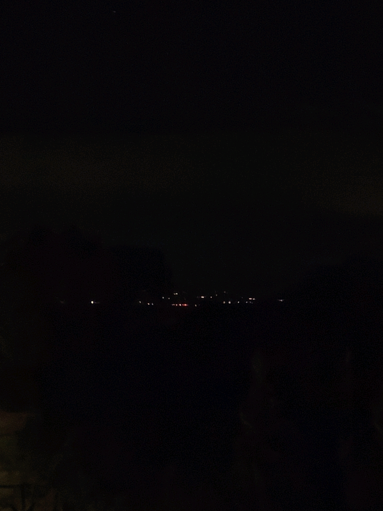This image depicts a dark night scene in Passignano sul Trasimeno, Italy. The sky is black, with a few faint lights visible in the distance, likely from buildings or streetlights. The foreground is dominated by deep shadows, making it difficult to discern any specific details of the environment or any potential subjects. There are no discernible people or objects in the frame. The overall impression is one of darkness and emptiness.