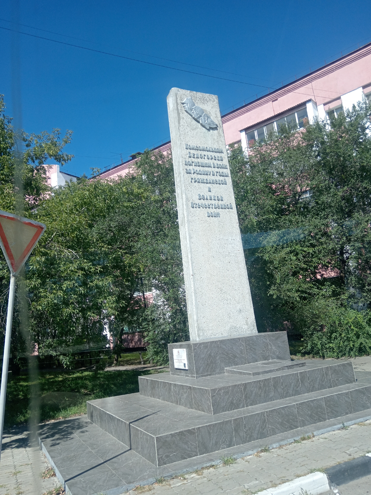 The image shows a memorial monument in the city of Belogorsk, Russia. The monument is a tall, grey obelisk with inscriptions and a relief on its surface. It stands on a multi-tiered base made of dark grey stone. Behind the monument, there are trees and a pink building with white trim and windows. In the foreground, there is a traffic sign, a sidewalk, and grass. The sky is clear and blue. The inscription on the monument appears to be in Russian and commemorates individuals who died in the Great Patriotic War.