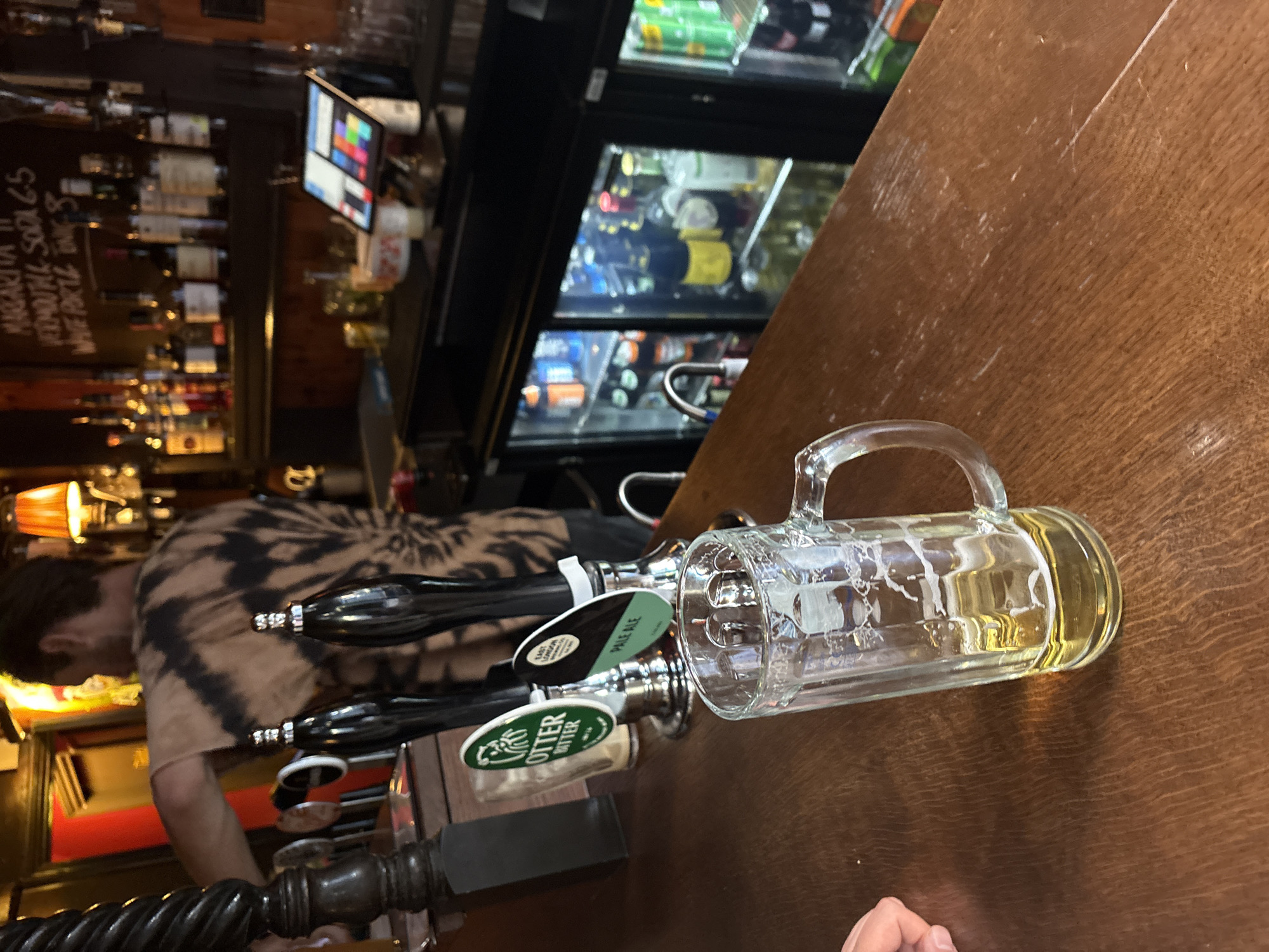 The image depicts the interior of a pub, potentially located in London, United Kingdom. At the forefront is a beer mug, partially filled with a golden liquid. Behind the mug are beer taps, labeled "Otter Bitter" and "Pale Ale," indicating a variety of local beers on tap. A man in a tie-dye shirt appears to be working behind the bar, potentially serving drinks or cleaning. The bar area includes a chilled display case filled with bottles and cans of beverages, and shelves stocked with various liquors are visible in the background. A chalkboard with writing is also discernible, possibly listing drink specials or prices. The overall ambiance suggests a traditional pub setting.