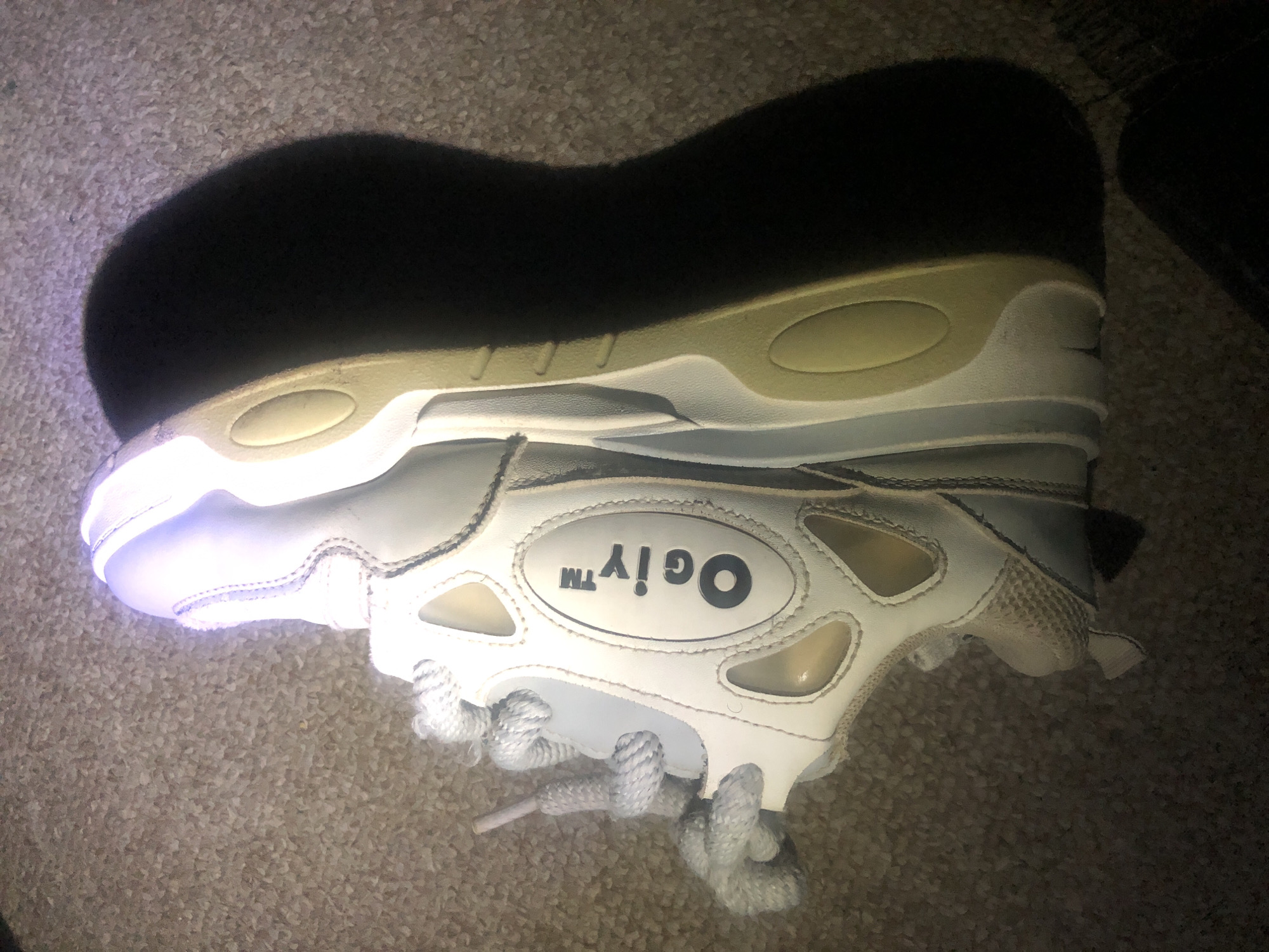 A single, white, laced athletic shoe with the brand name "Ogiytm" is lying on its side on a textured, brown carpet. The shoe is illuminated by a light source positioned above and to the left, casting a large, dark shadow to the right and partially onto the shoe's sole. The shoe appears to be made of leather and mesh, with prominent stitching and ventilation cutouts. The laces are thick and white. The sole of the shoe is thick and has a light tan color with some darker accents. The carpet is a light brown and appears to be loosely woven. No people or other objects are clearly visible in the image. The scene is static. There are no discernible features that indicate a specific location within the city of Jalingo, Nigeria.